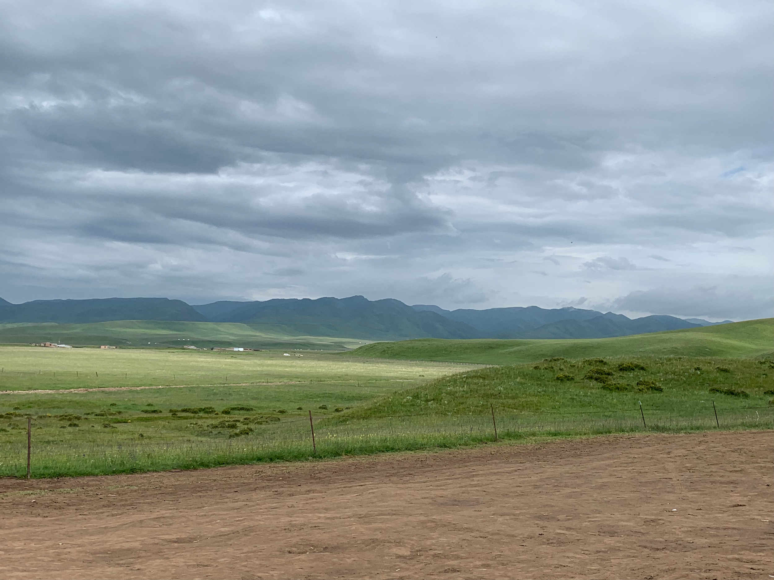 A wide view of a green landscape with rolling hills, mountains in the distance, and a cloudy sky overhead, with a dirt road in the foreground.