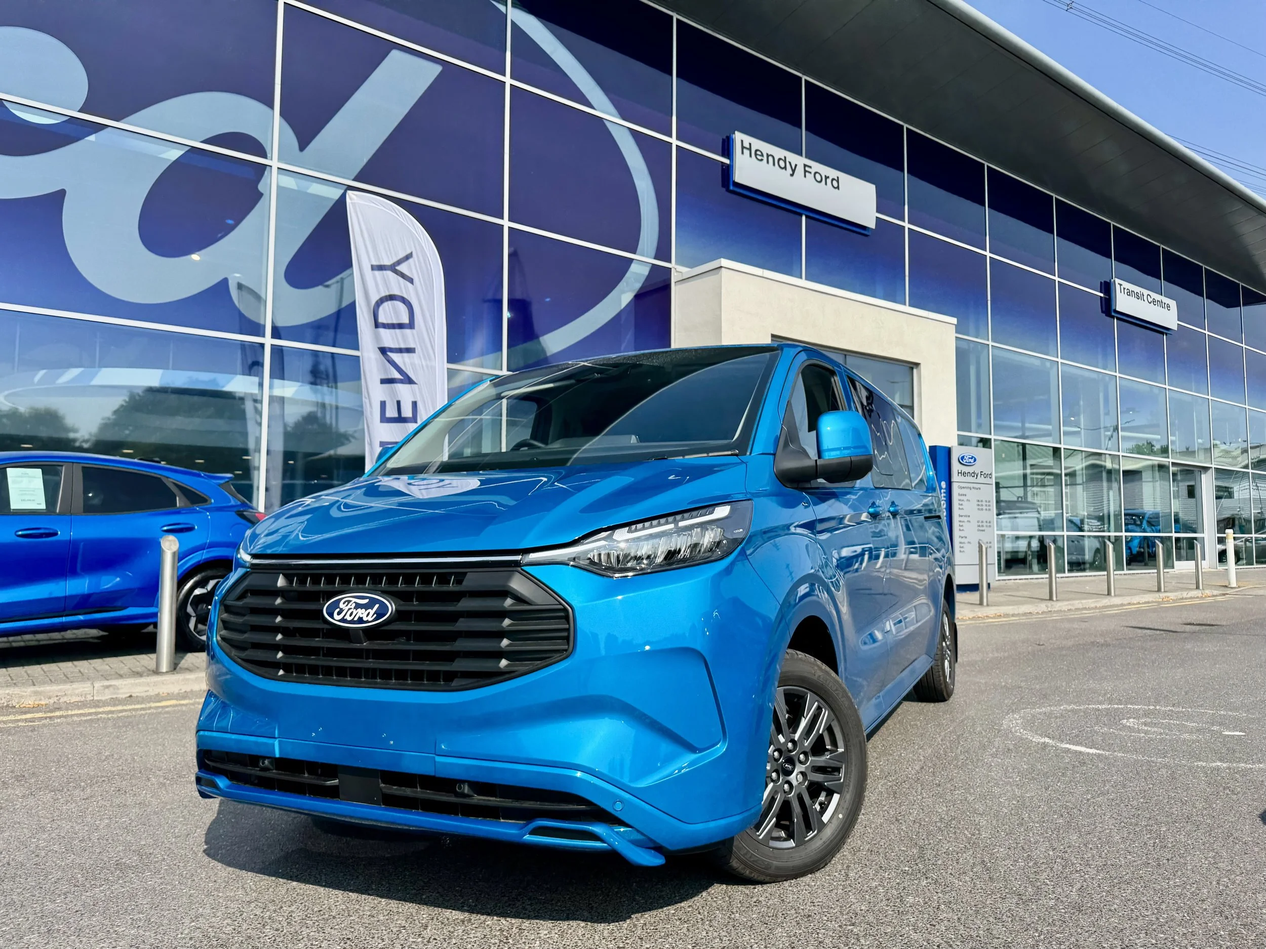 Blue Ford Transit custom Double Cab Sport van parked outside Hendy Ford dealership, with glass building featuring Ford signage and a flag with Ford logo in the background. the vehicle is fitted with a Sports body kit. 