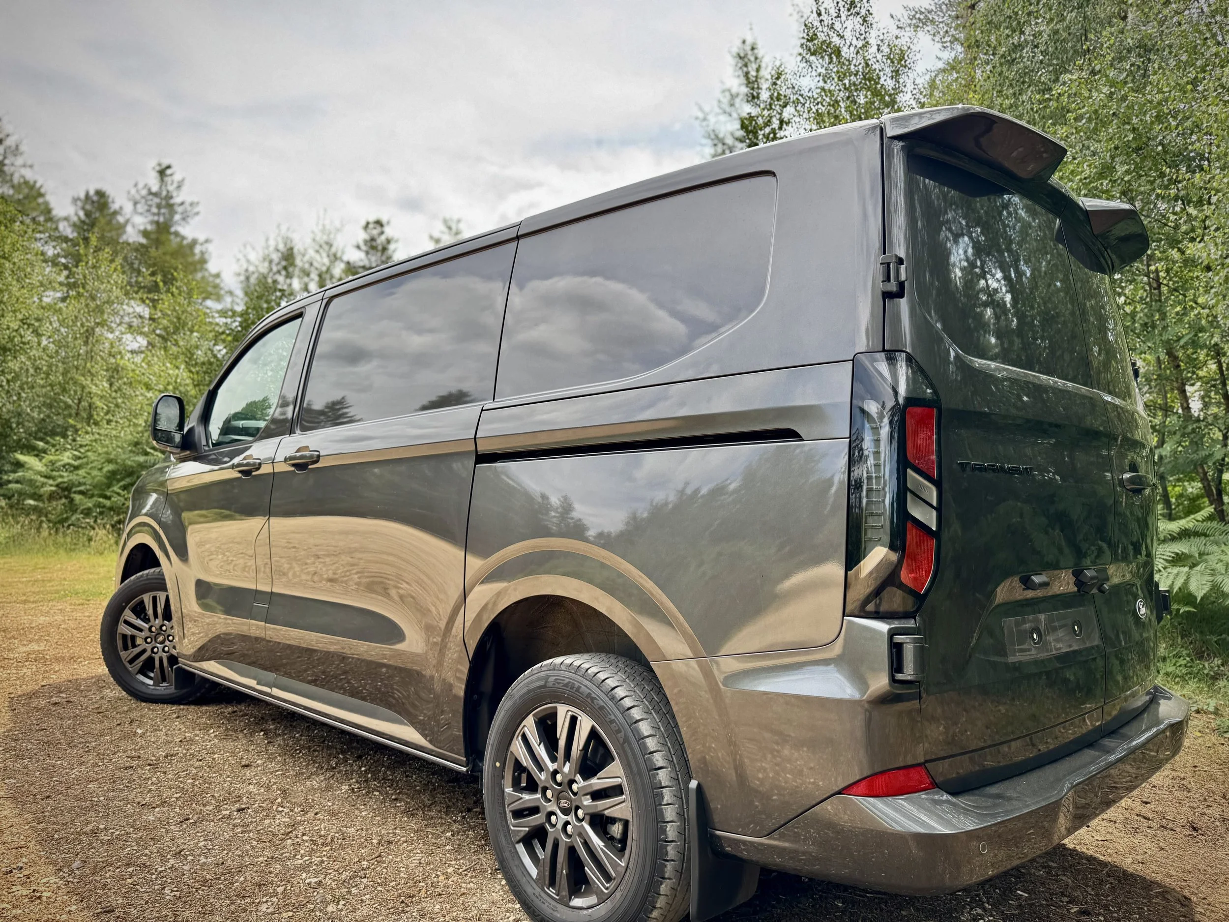 Gray Ford Transit van parked on a gravel area with green trees and a cloudy sky in the background.