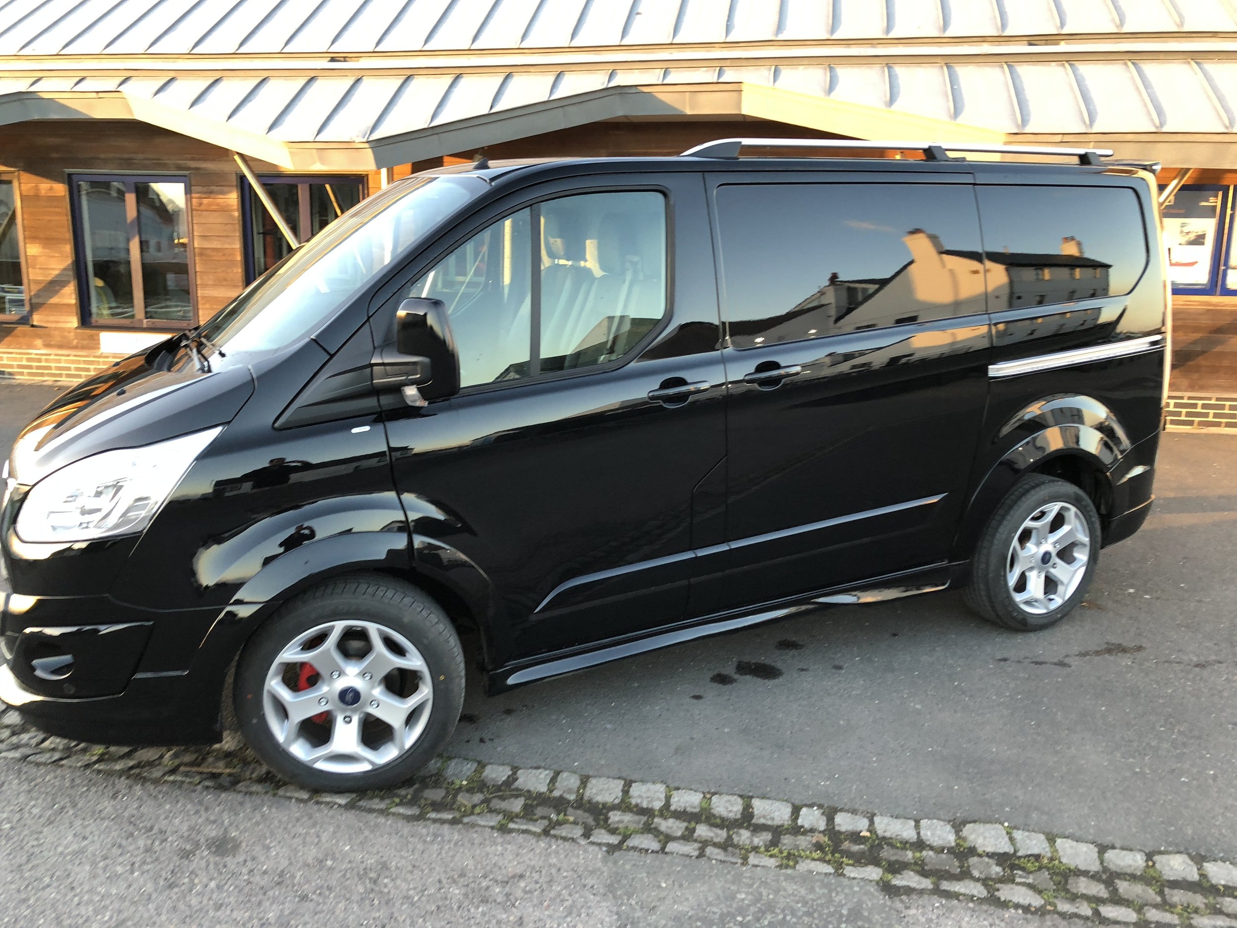 Black Ford Custom Panel van parked on a street in front of a building with wooden walls and large windows. the. van has an SV Sport body kit installed, with side skirts and silver alloy wheels 