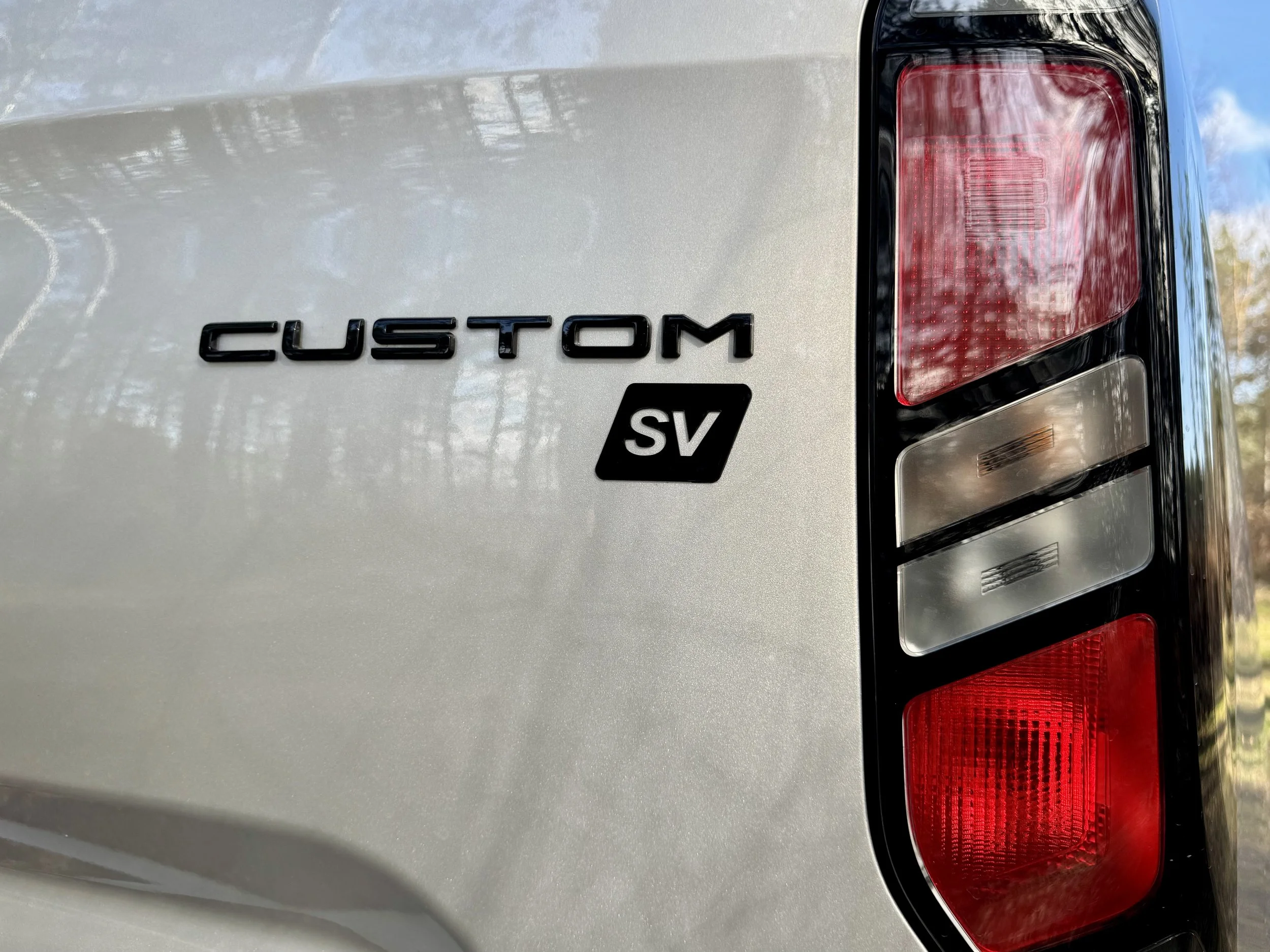 Close-up of the rear of a silver vehicle showing the black 'CUSTOM' badge and black 'SV' badge on the tailgate, with part of the taillight visible.