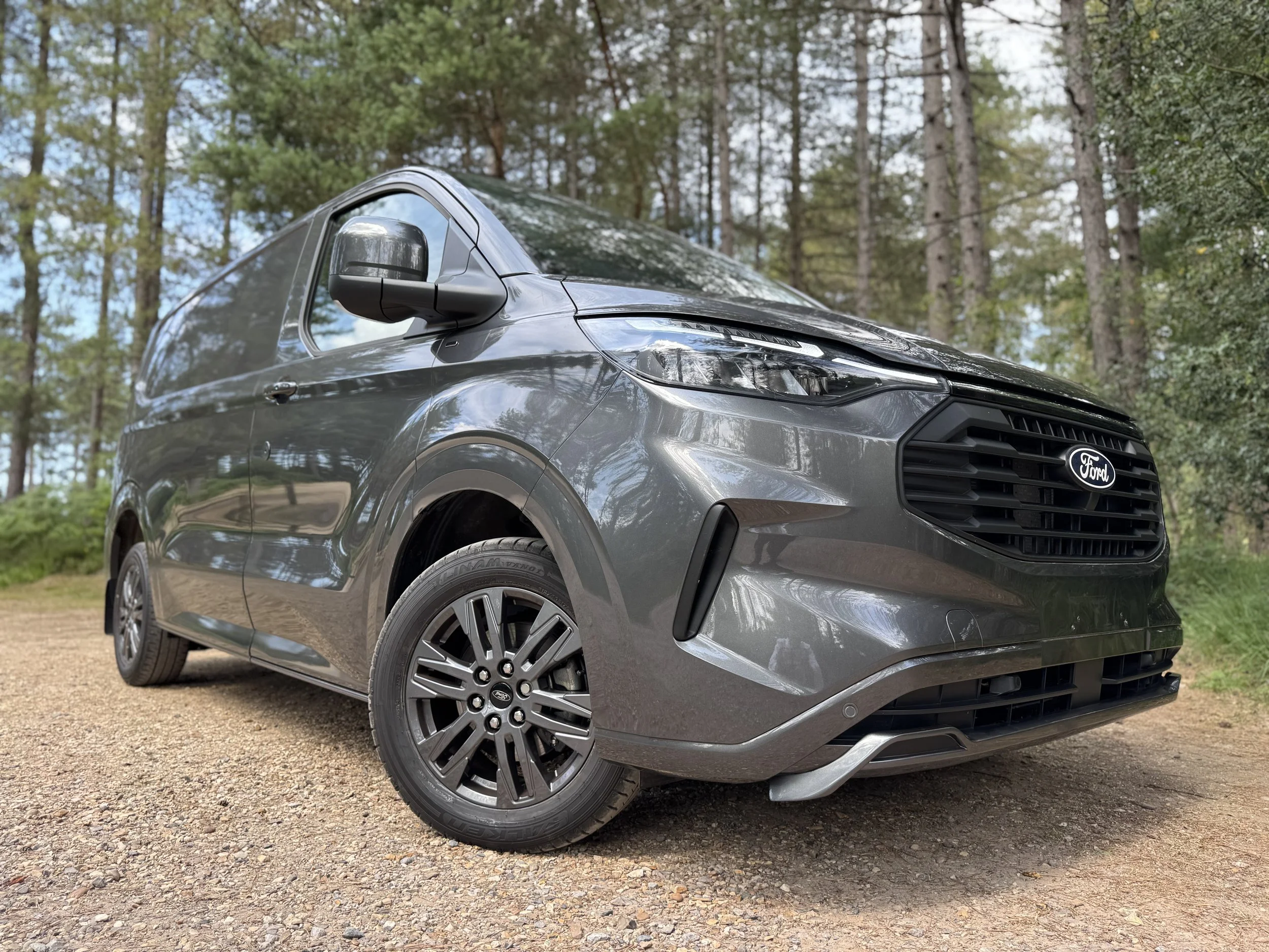 A gray Ford Transit Panel van - S Sport,  parked on a dirt path in a forested area with tall trees and green foliage.