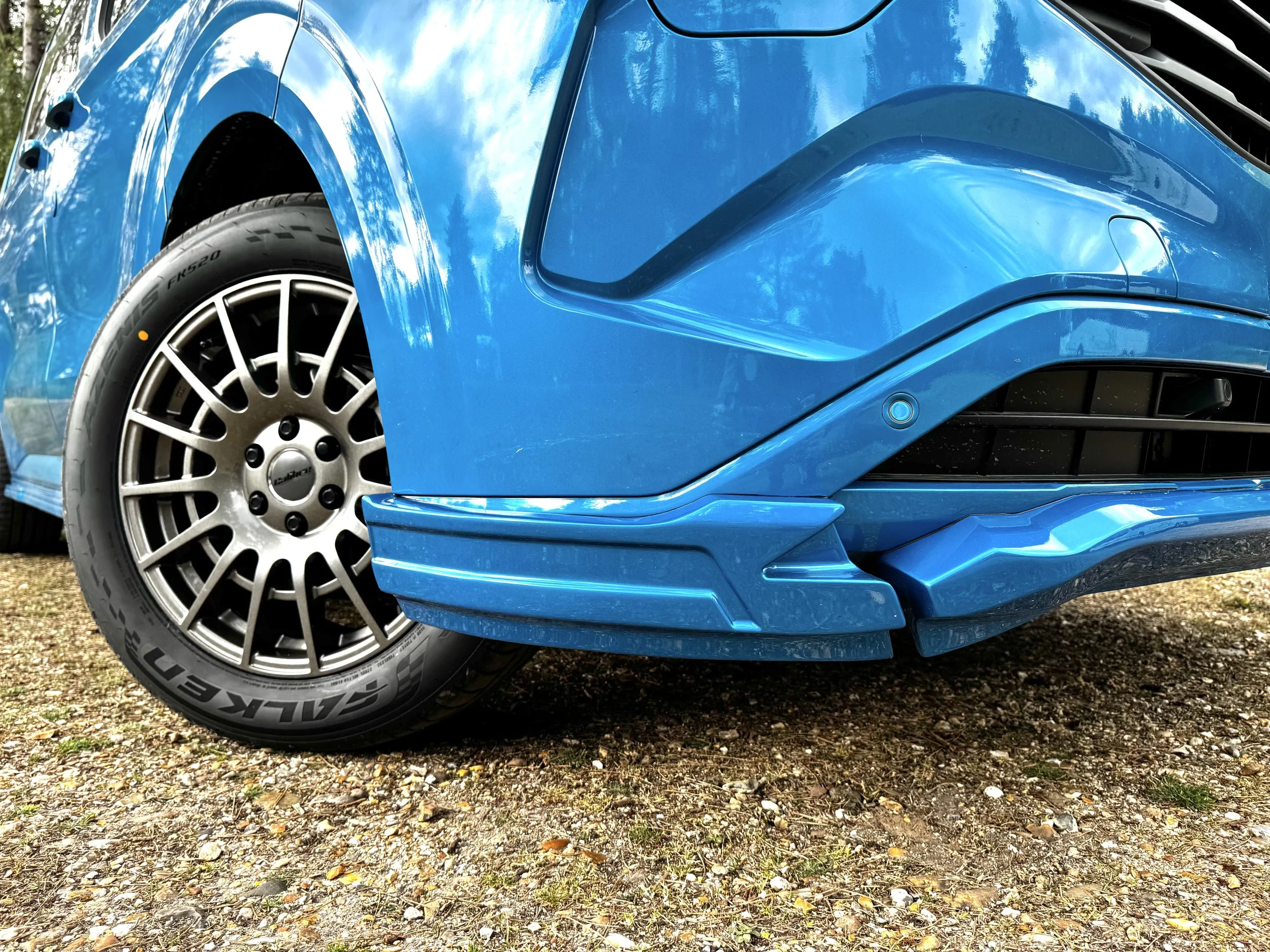 Picture of a front bumper of a Ford Custom SV Sport Crew van in digital blue. in a forest landscape background