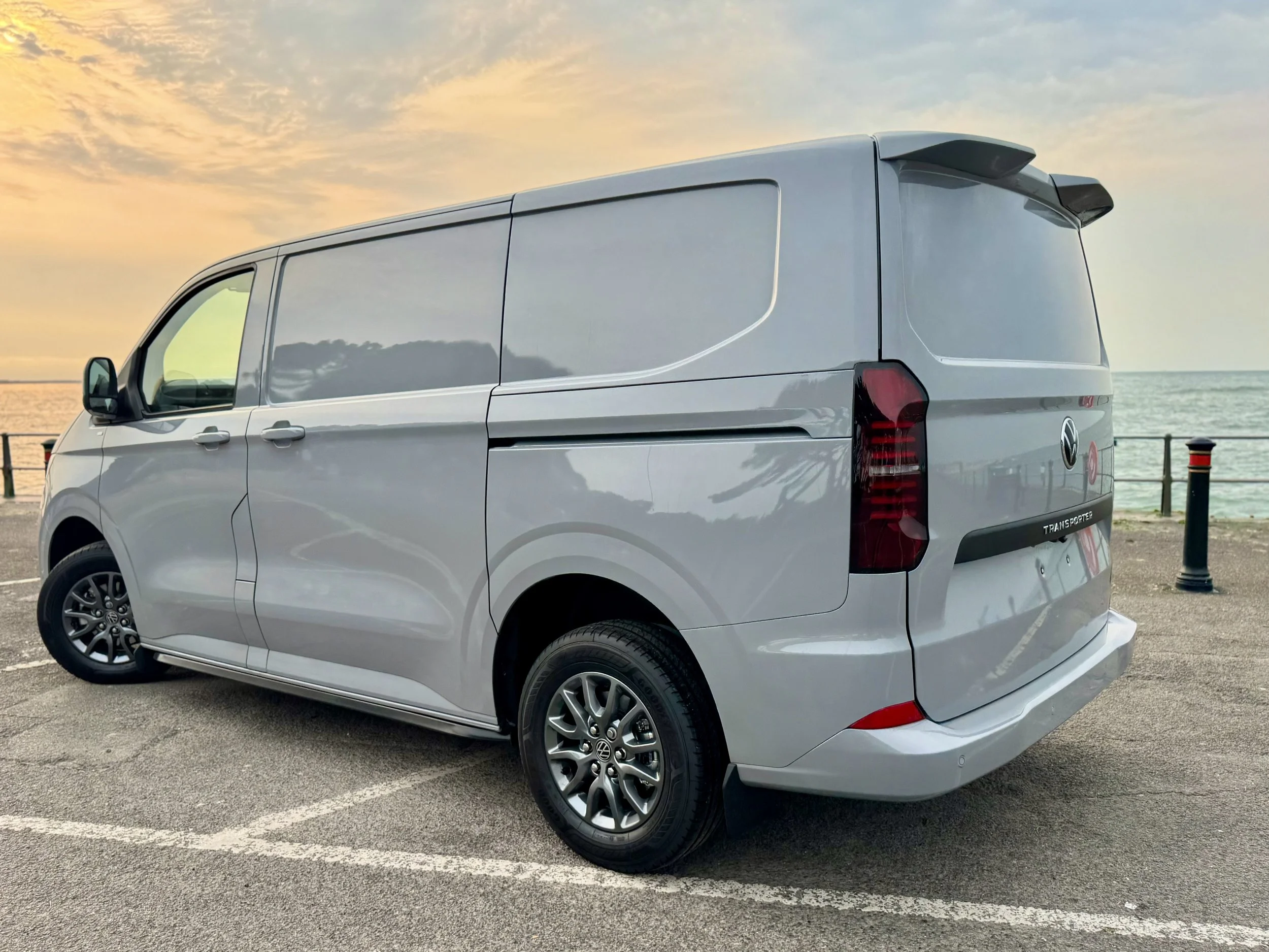 A silver Volkswagen Transporter van parked in a lot near the beach during sunset.