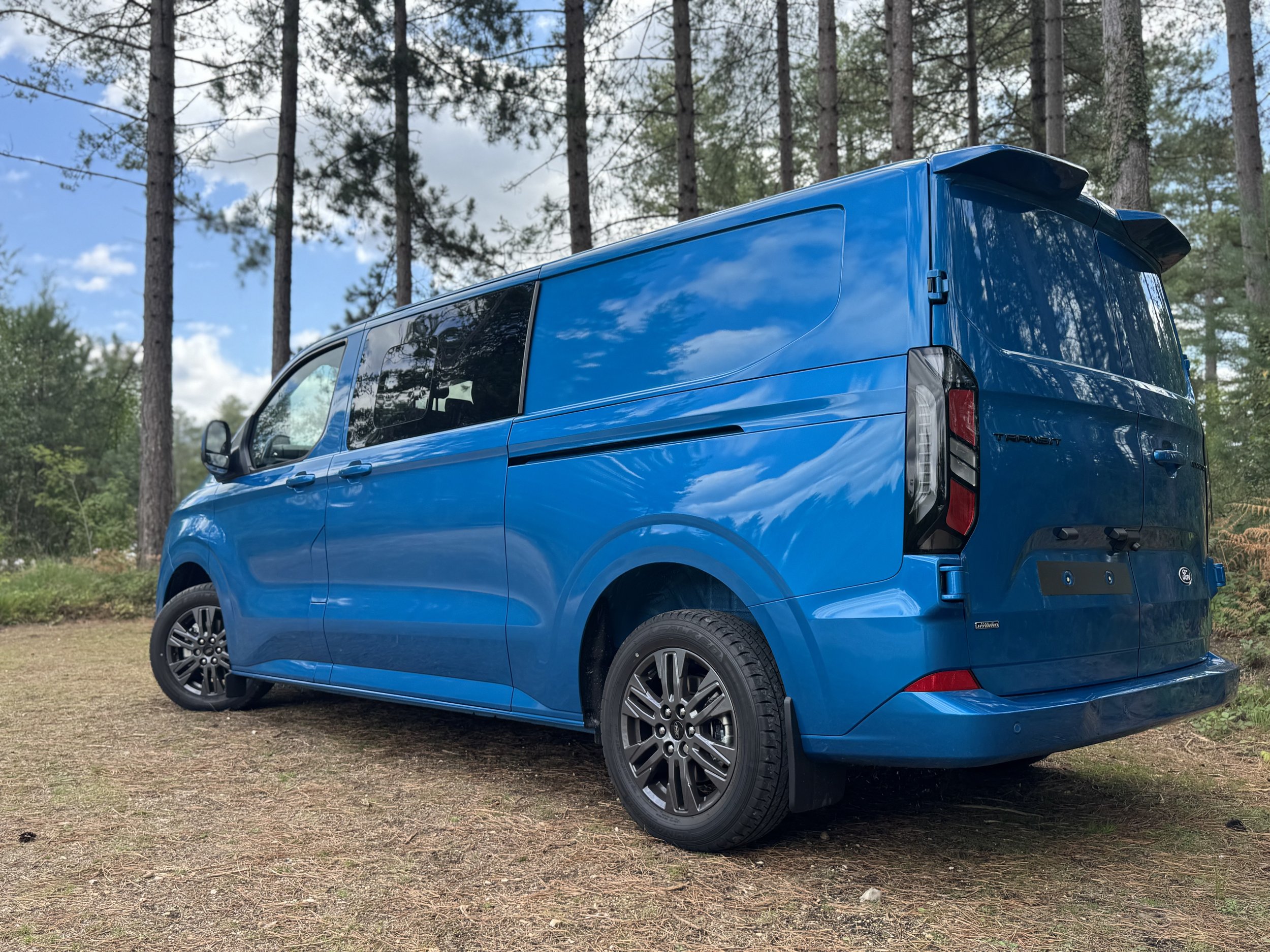 Blue Ford Transit Custom Sport Van,  parked on dirt in front of a forest of tall trees with a partly cloudy sky.
