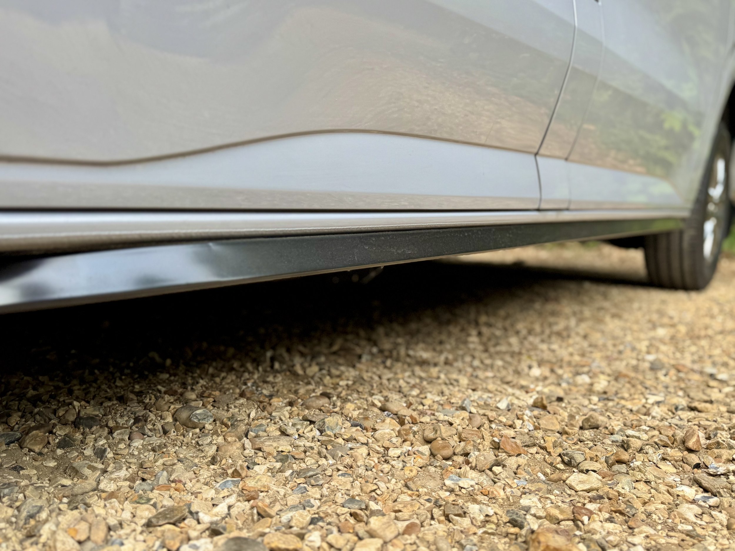 Close-up of a silver car's side skirt and black running board, parked on gravel ground, with part of the rear tire visible.