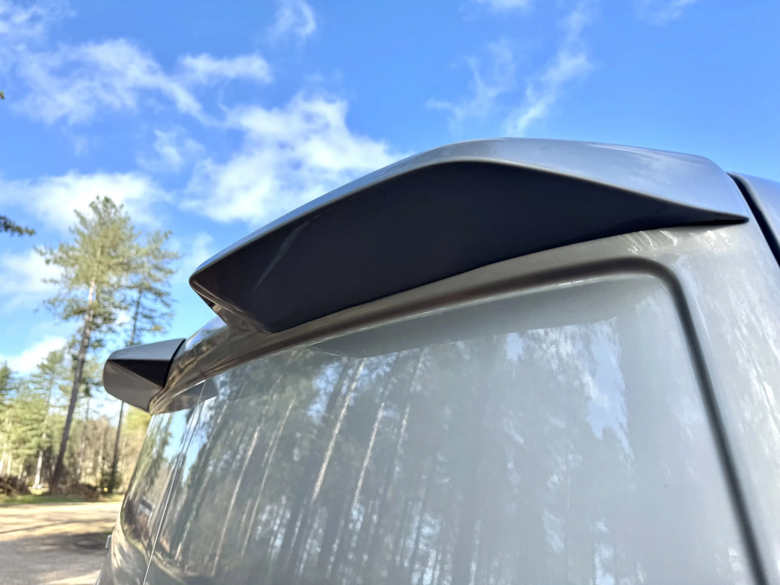 Close-up of the rear roof spoiler of a silver or light gray vehicle with a clear blue sky and trees in the background.