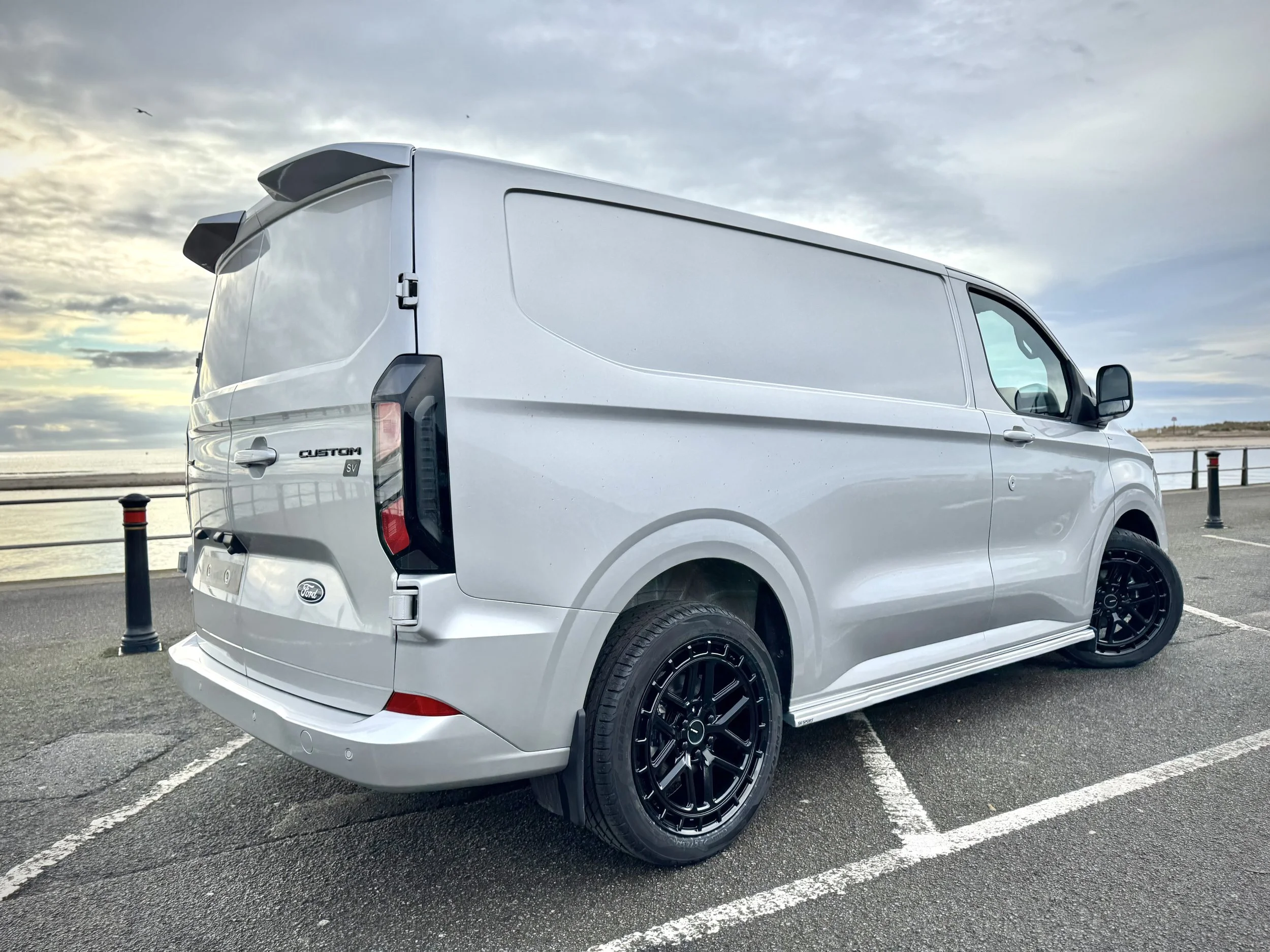 White Ford Custom van parked on a seaside parking lot during cloudy weather, with black wheels and tinted rear windows.
