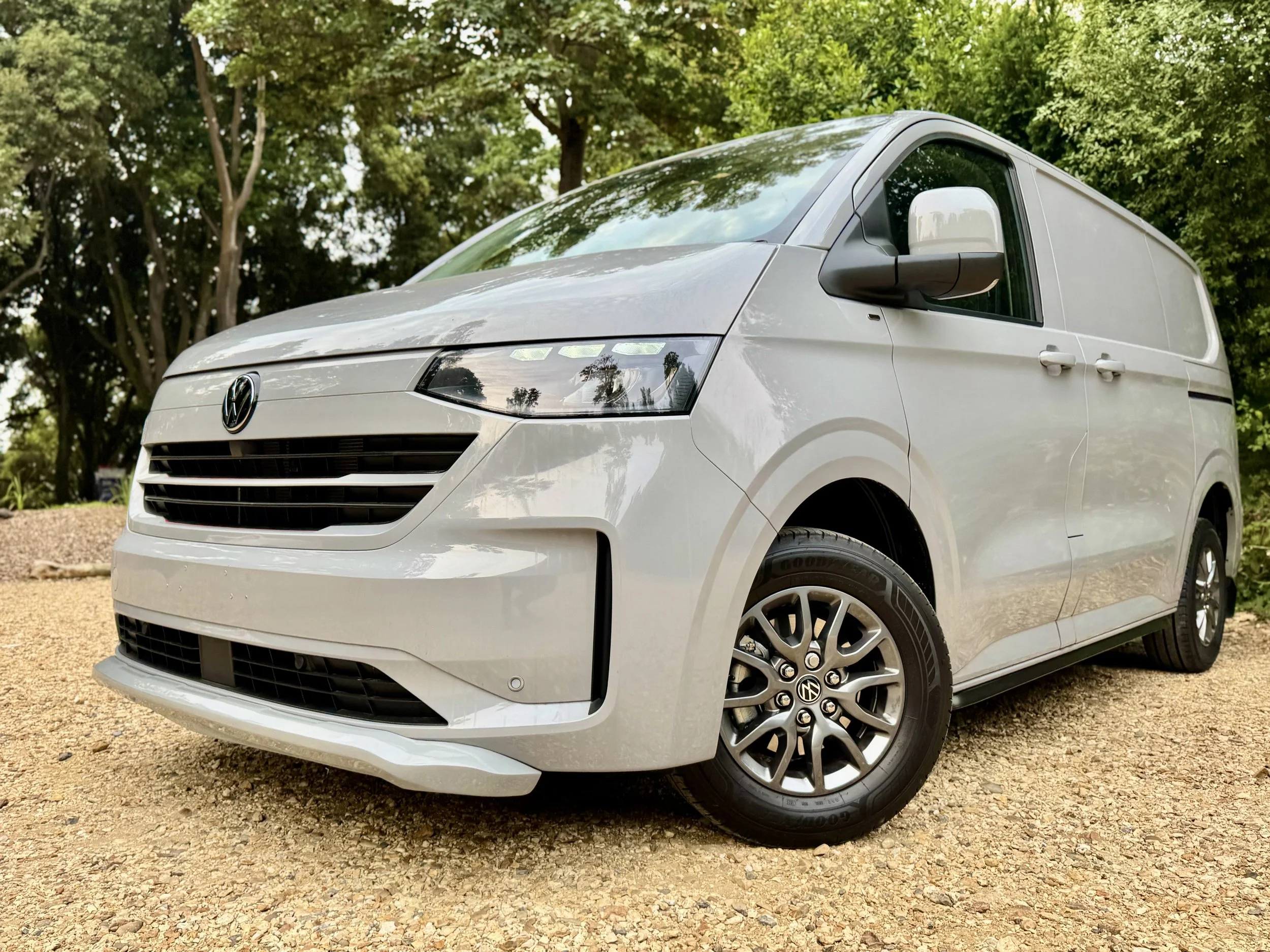 White Volkswagen delivery van parked on gravel ground with green trees in background.