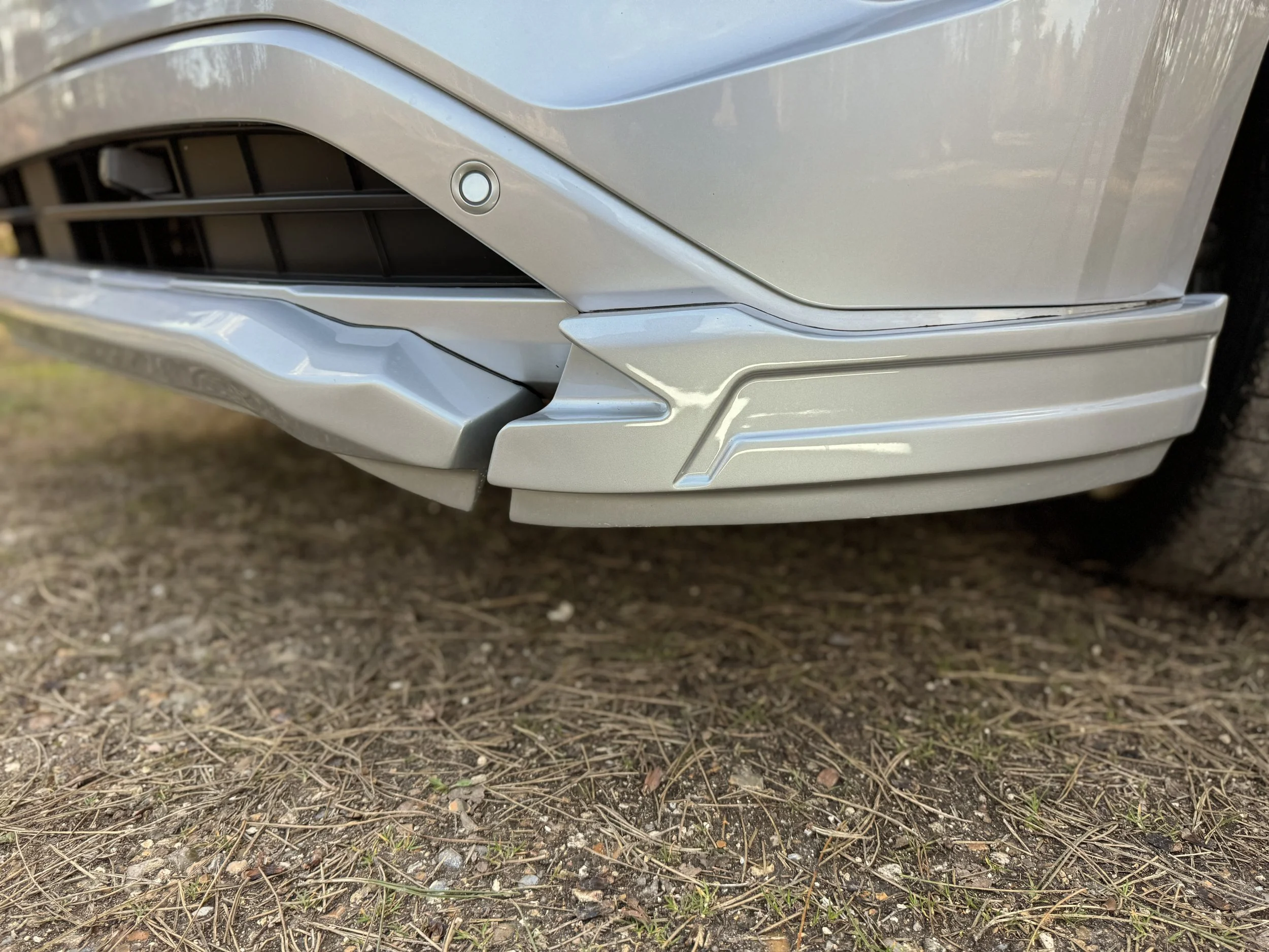 Close-up of the front lower bumper and grille area of a silver or light gray car parked on dirt or gravel ground.