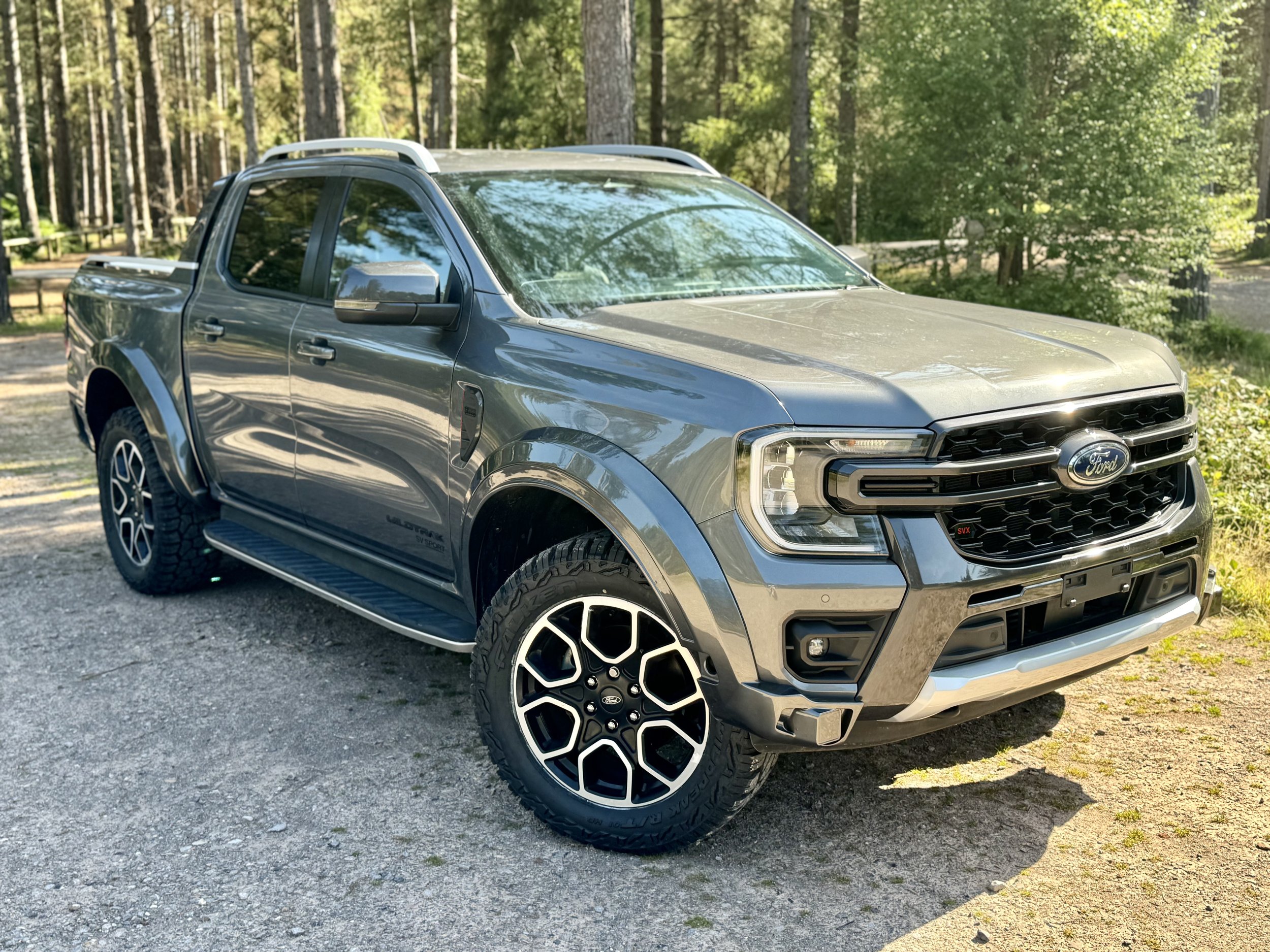 Gray Ford pickup truck parked on a gravel surface in a wooded area.