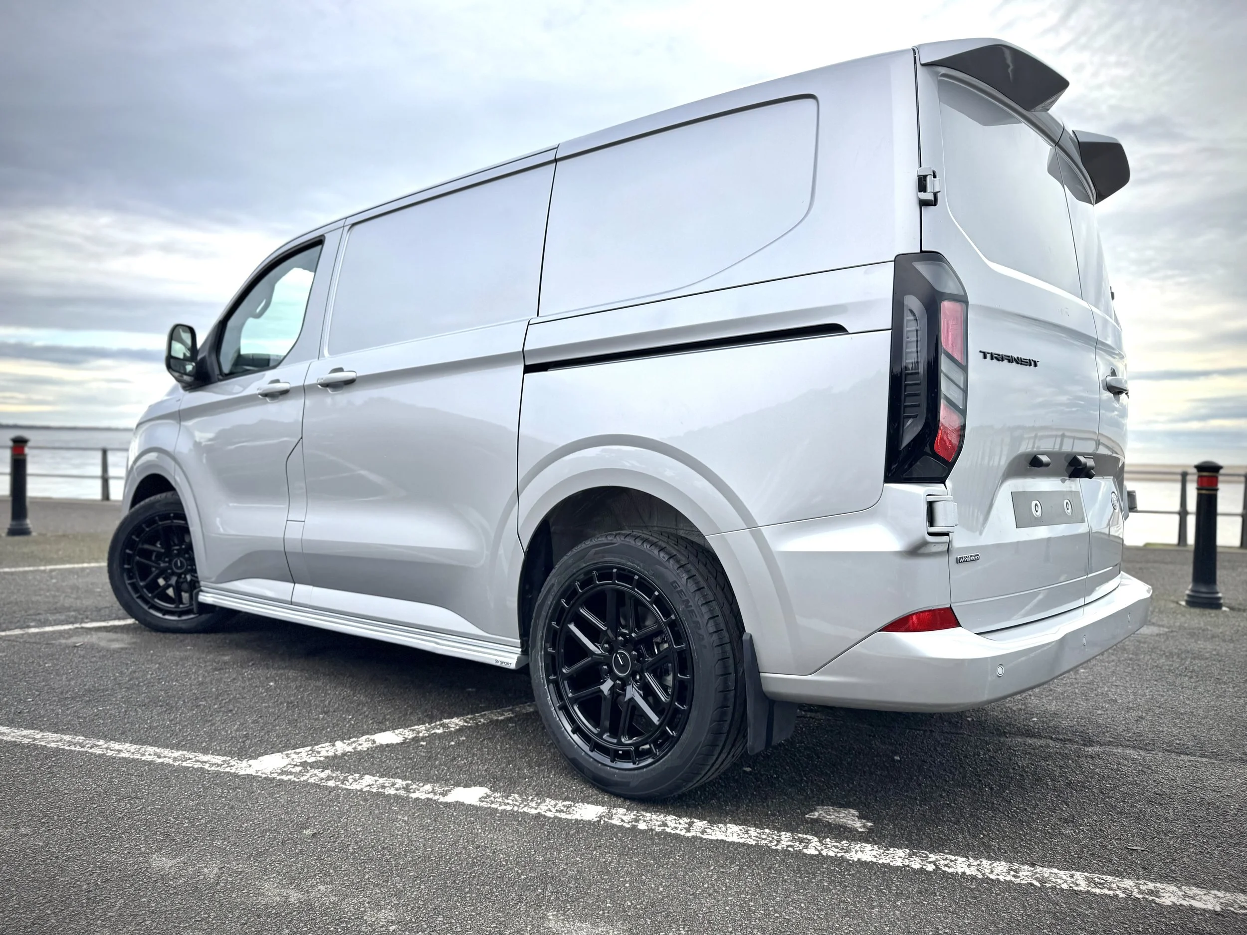 A white Ford Transit van parked at a waterfront parking lot with black custom wheels and a cloudy sky.
