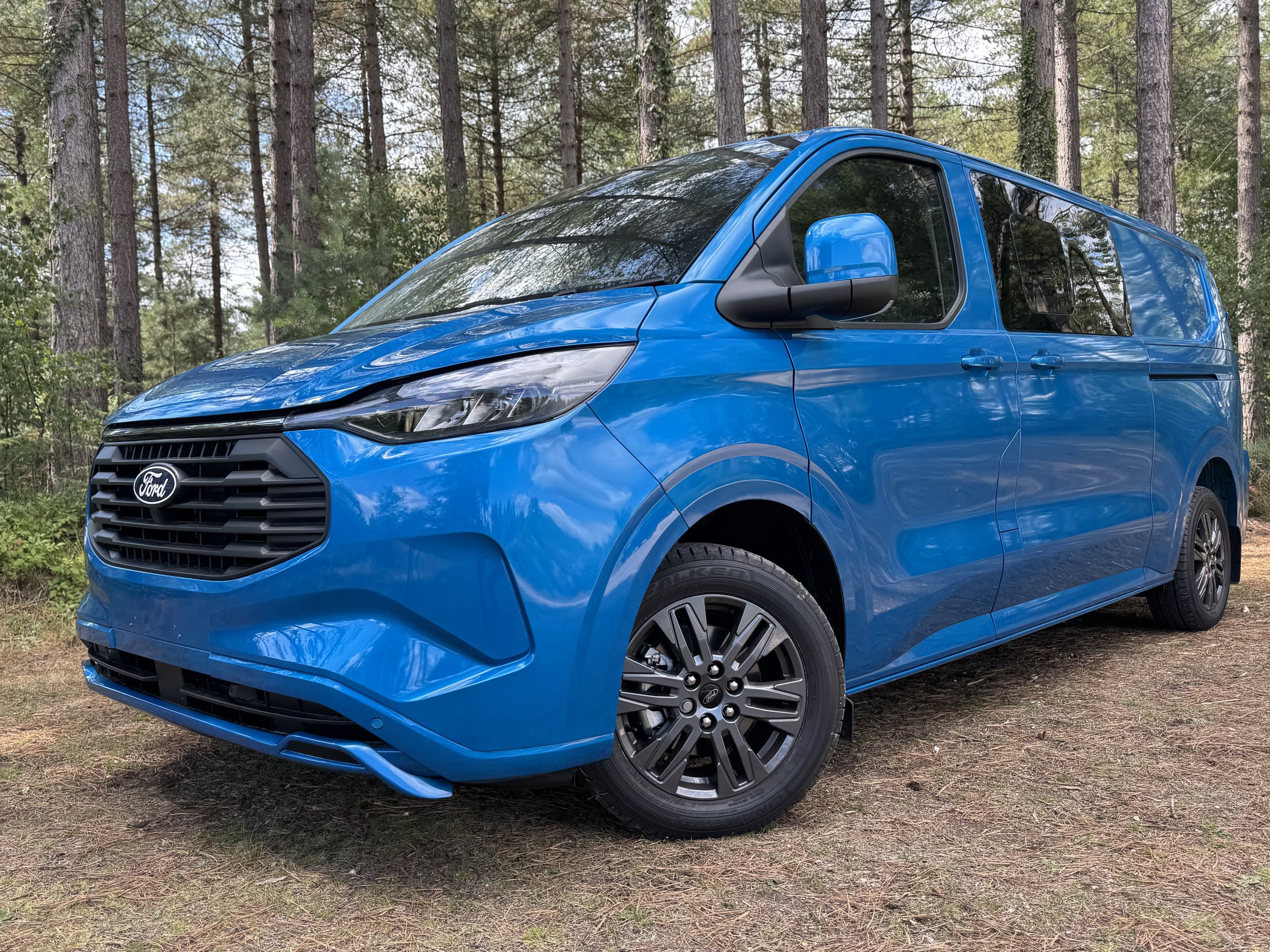 Blue Ford Transit Connect van parked on a dirt ground in a forest with tall trees.