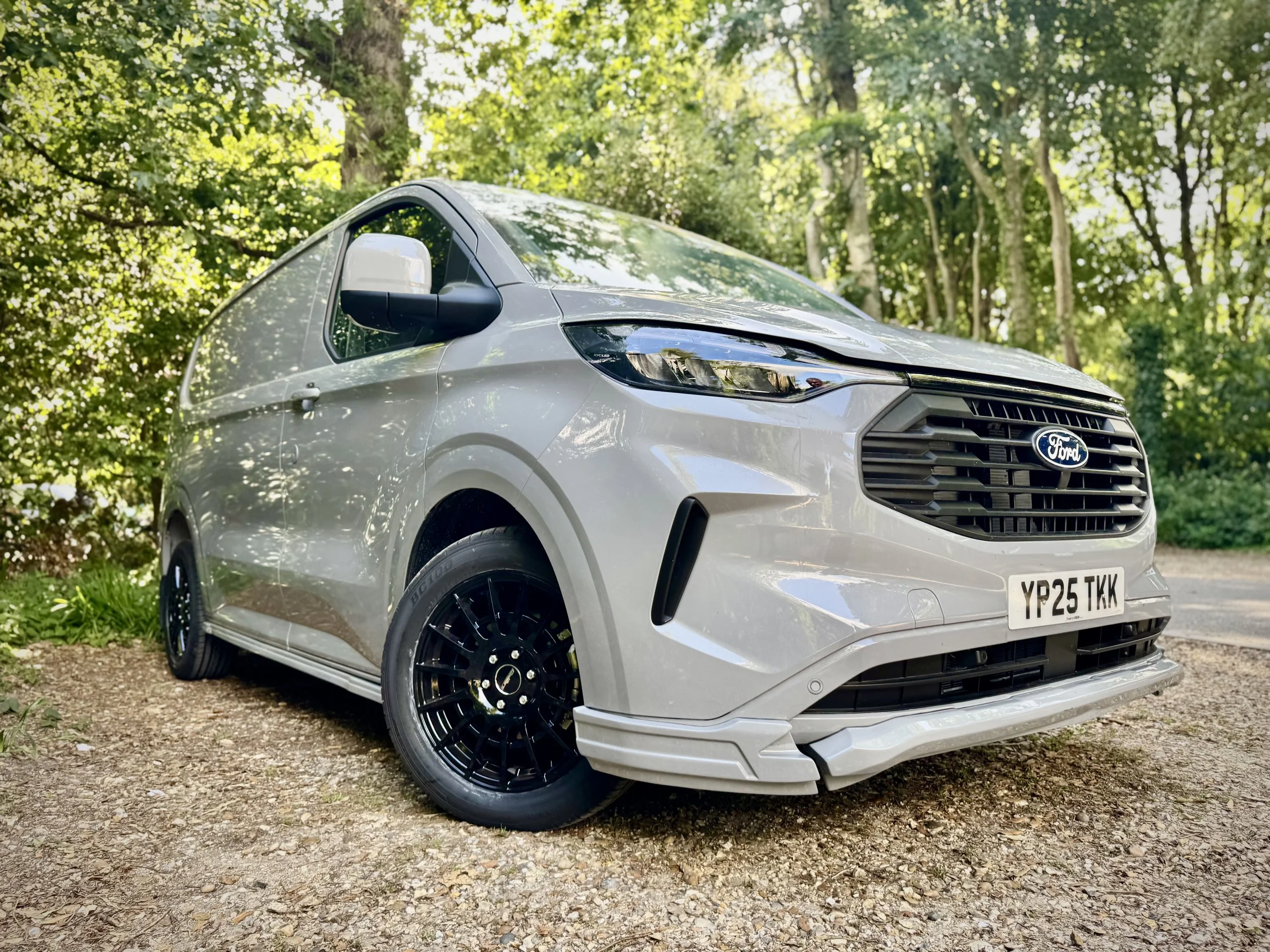 A silver Ford Transit Custom SV Sport van parked on a gravel surface with a forested background.