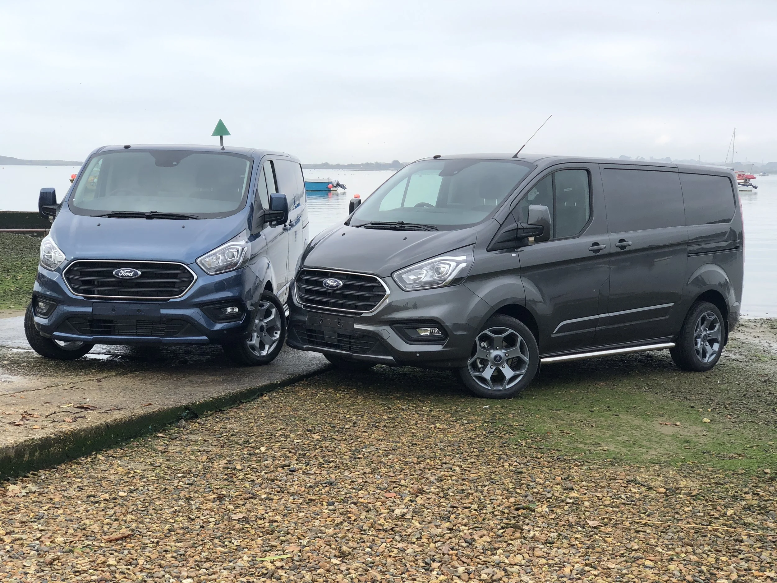 Two Ford Custom S Sport vans from SV Sport, one blue and one grey, parked by the water on a pebbled surface with a cloudy sky and boats in the background.
