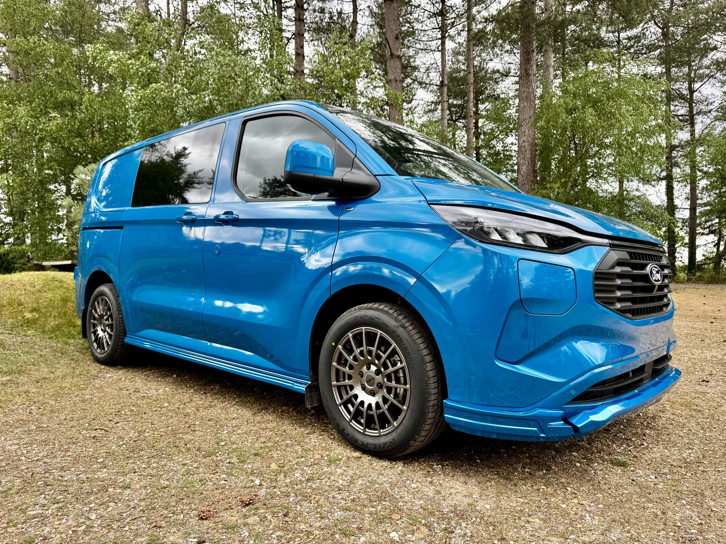 Image of a ford custom crew van in a digital blue paint, it has the SVX sports kit from SV Sport Vans fitted giving it a sporty look, the photo is in a forest setting in summer