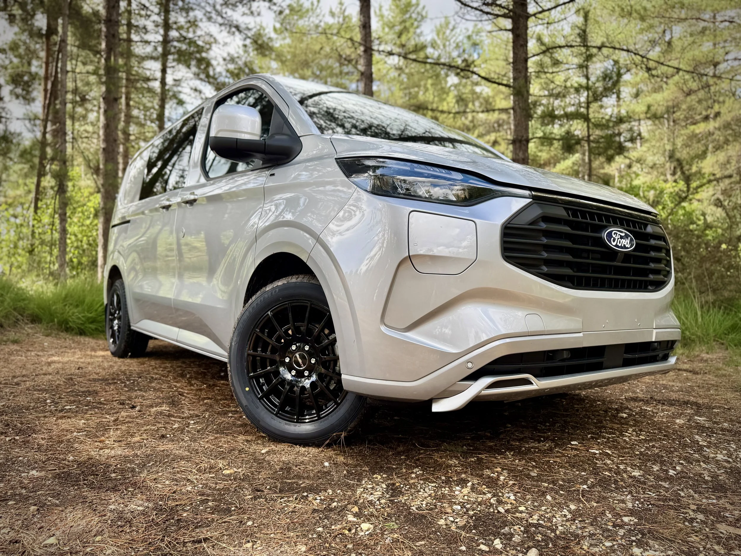 Silver Ford van parked on dirt in a wooded area with trees and green foliage in the background. The vehicles is a Custom Crew Cab and has a body kit fitted and black  18 inch alloy wheels