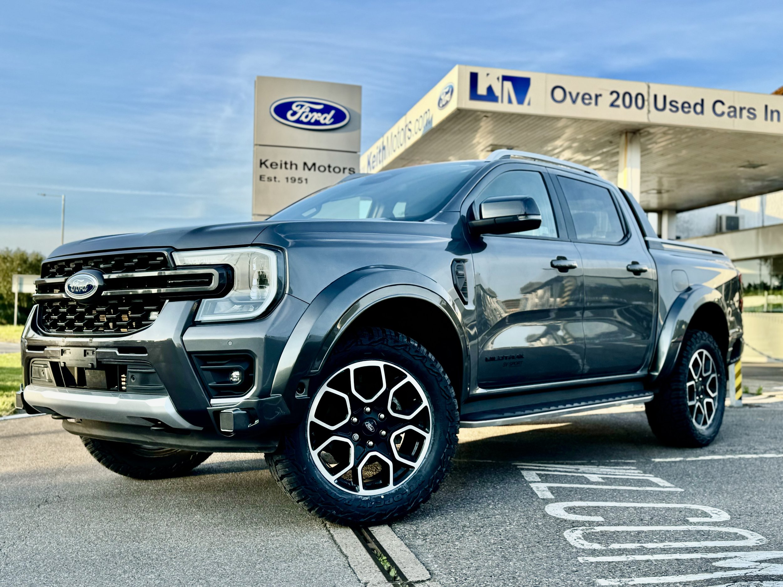 A gray Ford pickup truck parked in front of a used car dealership with a Ford sign and a building advertising over 200 used cars.
