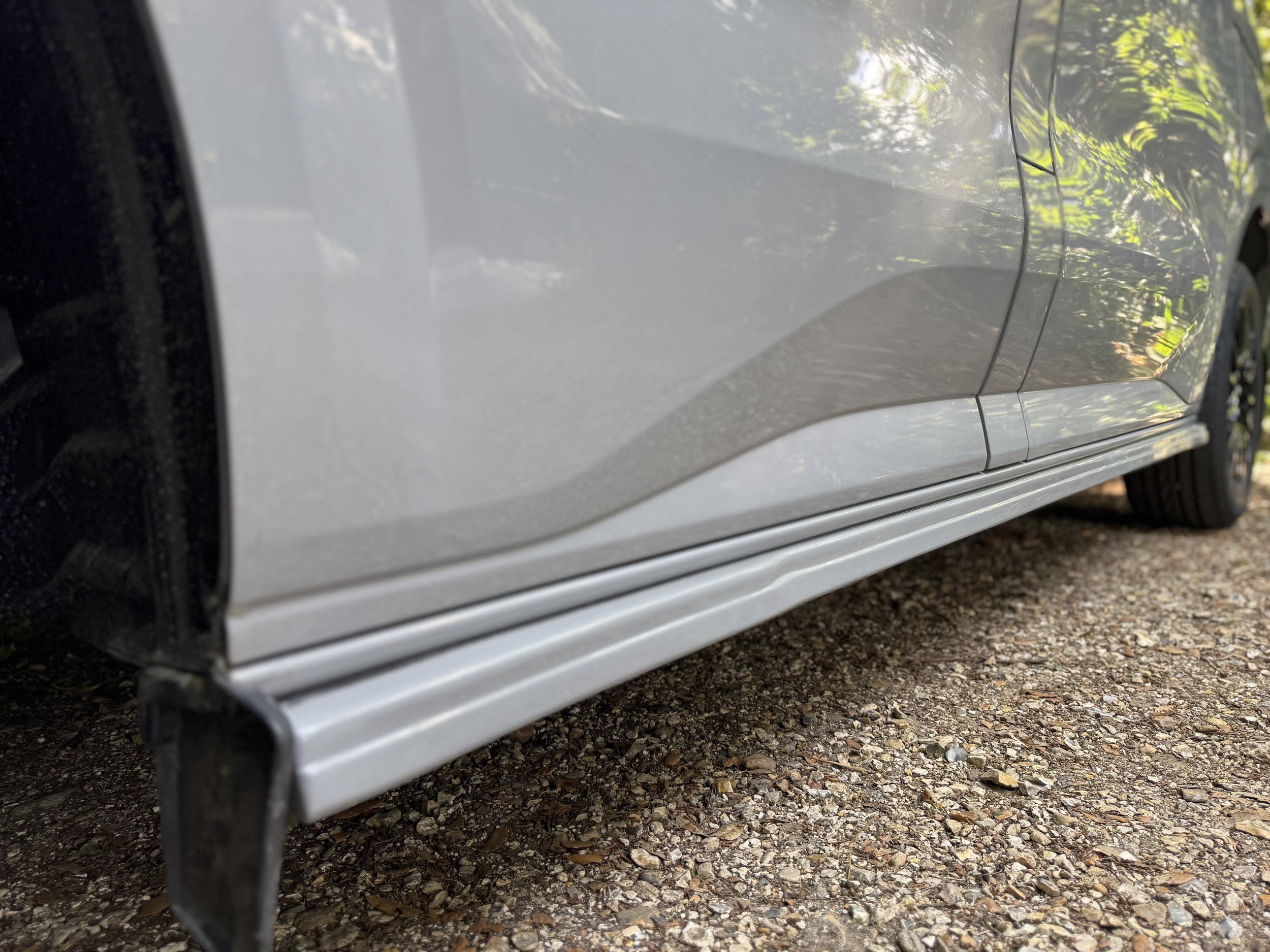 Close-up of a silver vehicle's lower side, showing the side skirt and part of the wheel on a gravel surface with trees reflected on the car's surface.