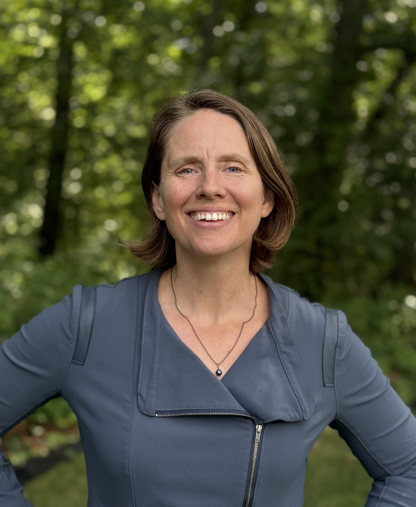 An image of Sarah Whiting, candidate for School Committee. She has short brown hair and is wearing a jacket, standing in front of some trees.