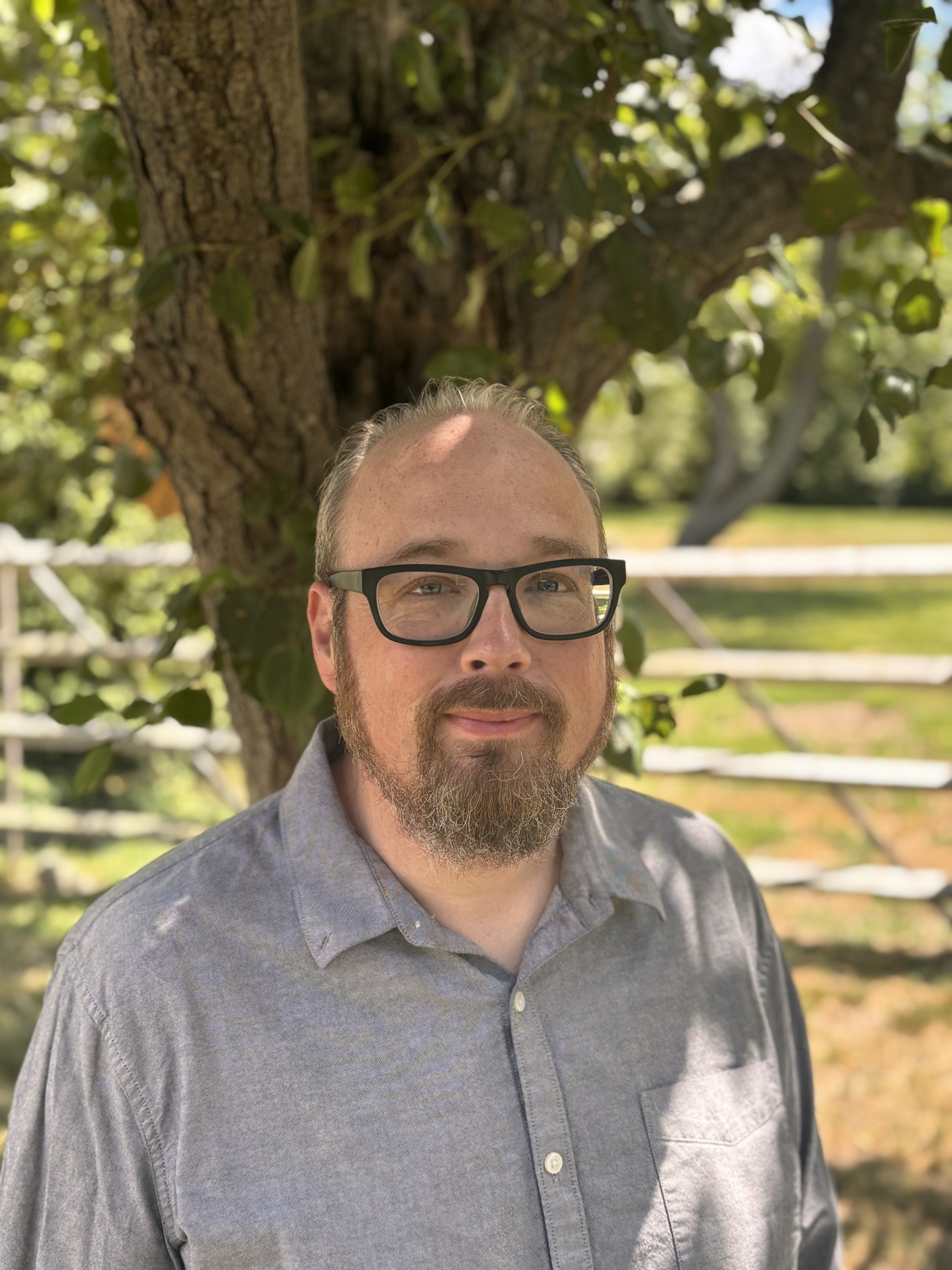 An image of School Committee candidate Adam Stearnbourne. He is wearing a gray button-up shirt and black glasses, and standing in front of a tree.
