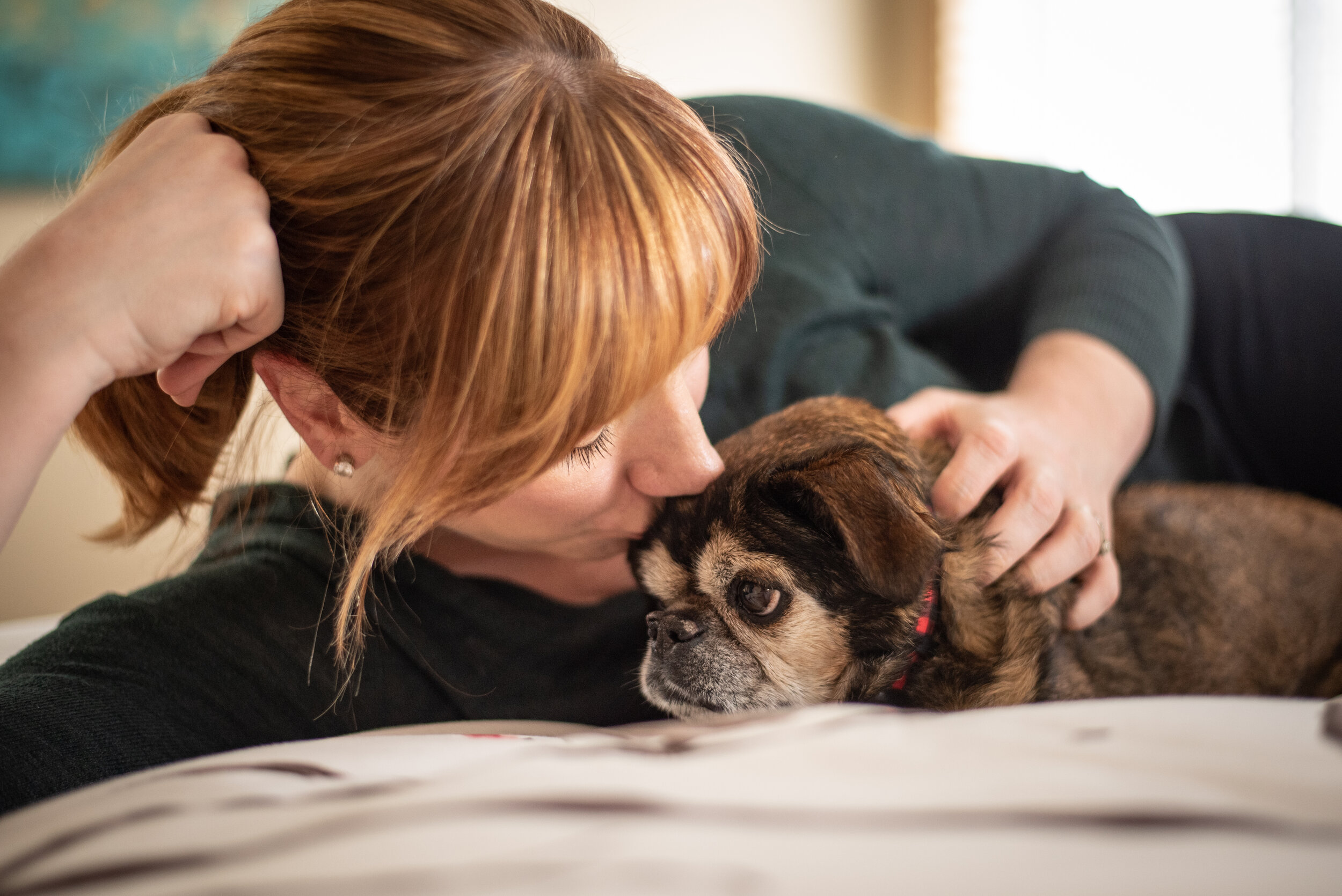 A small dog on the bed getting a kiss from a women with a ponytail