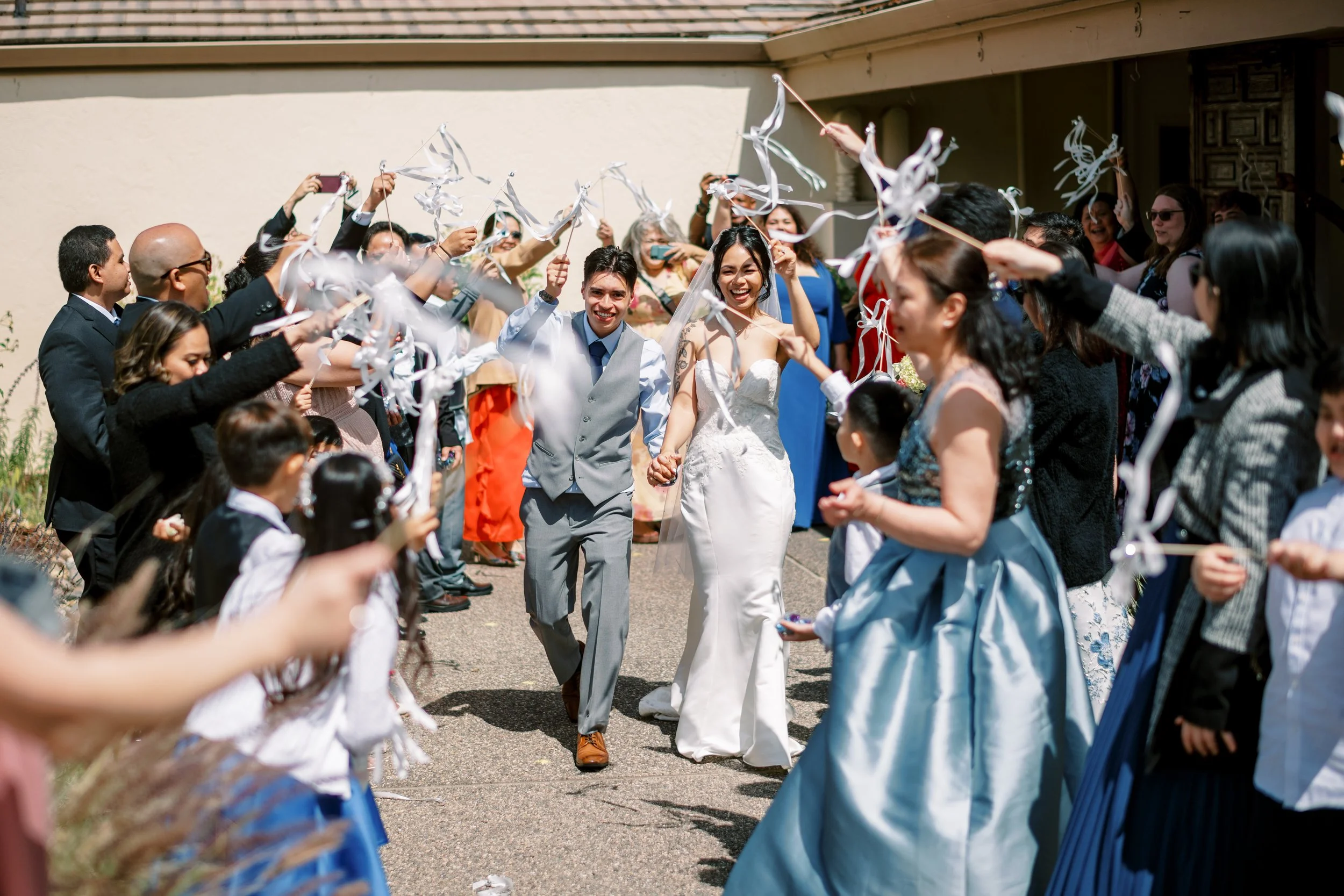 Bride and groom walking hand-in-hand through a crowd of wedding guests holding ribbon wands and celebrating outside on a sunny day.