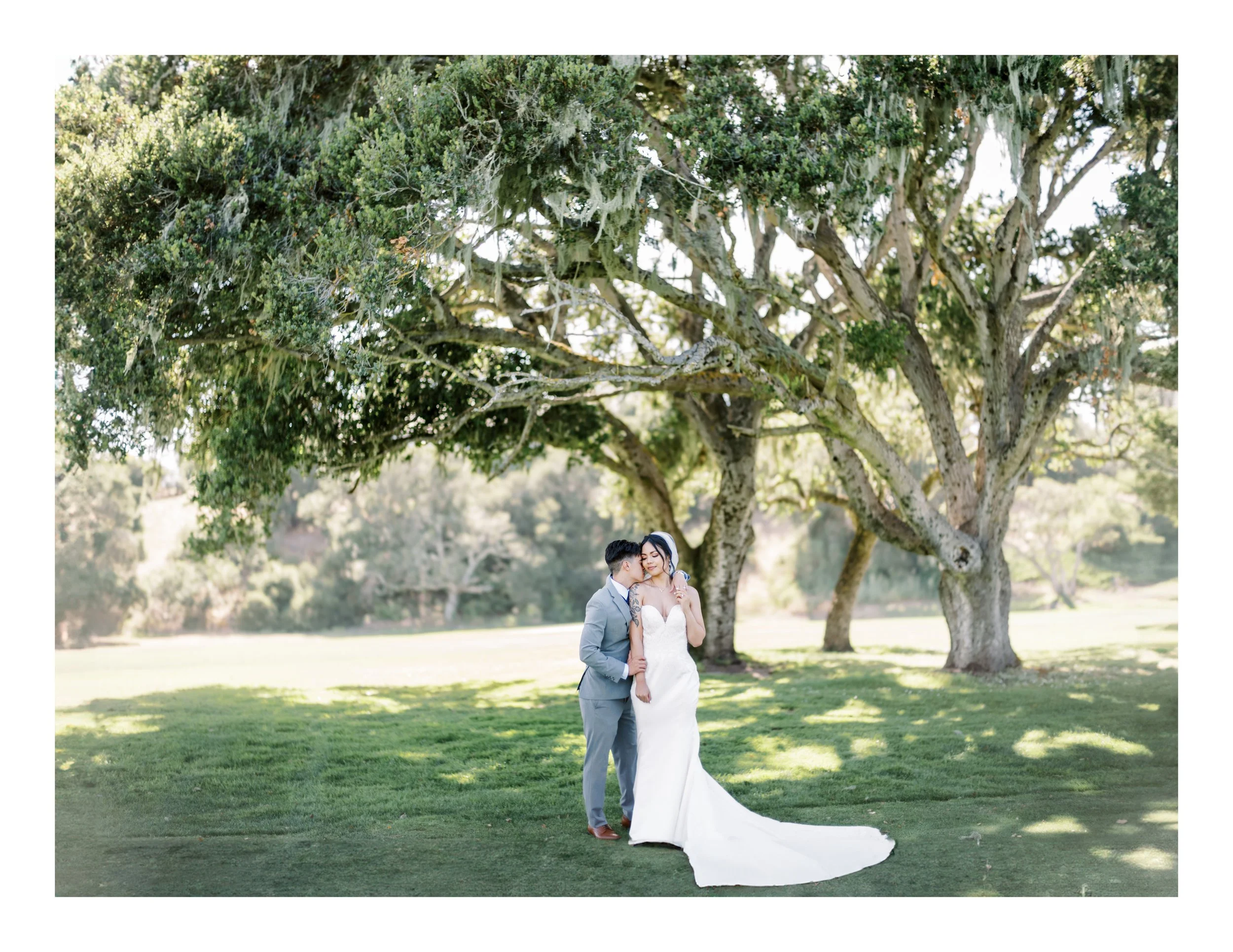 A bride and groom stand together under a large, leafy tree on a grassy lawn in a park setting, dressed in wedding attire.