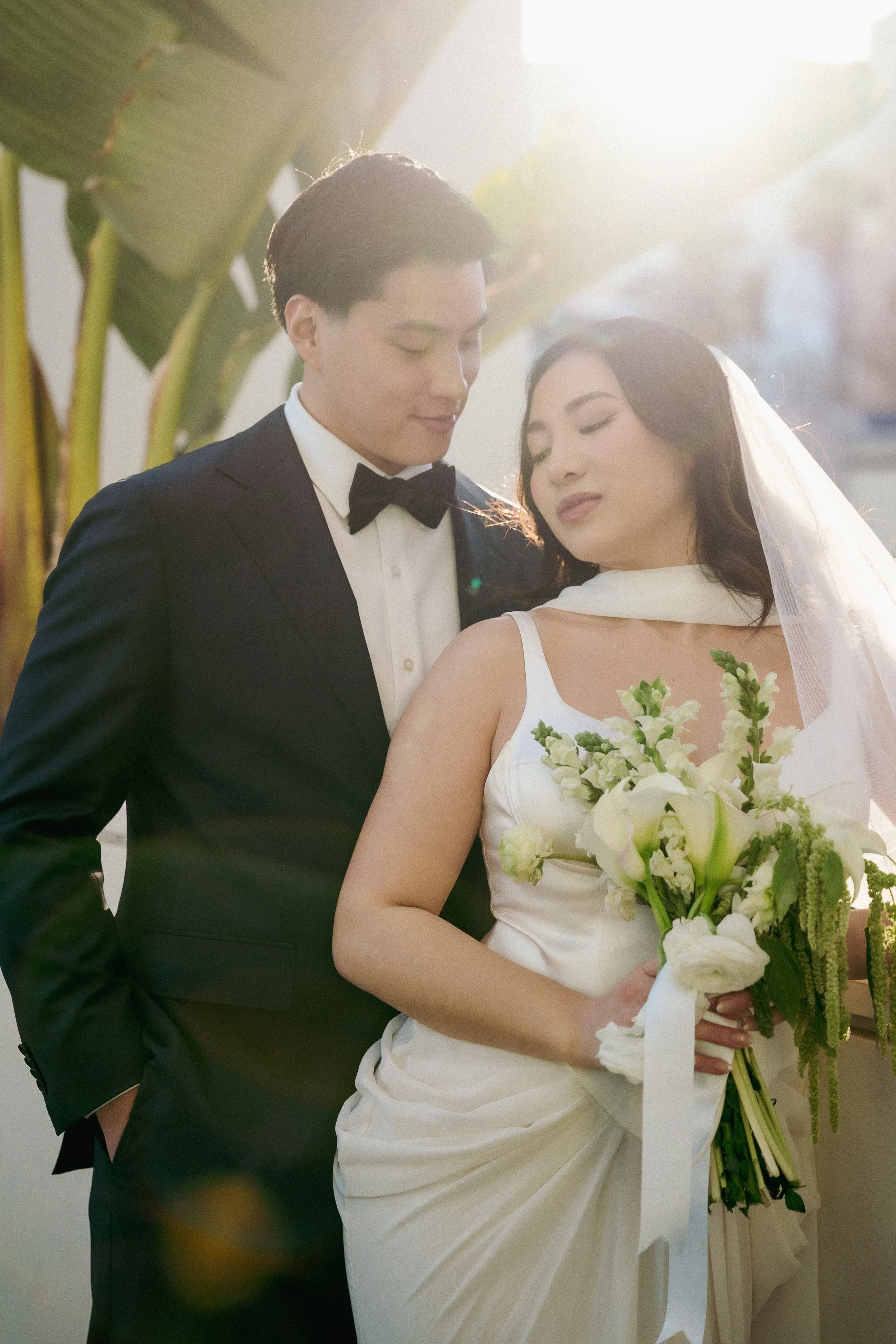 A bride and groom on their wedding day, standing close with their heads tilted towards each other, the bride holding a bouquet of white flowers, with sunlight in the background.