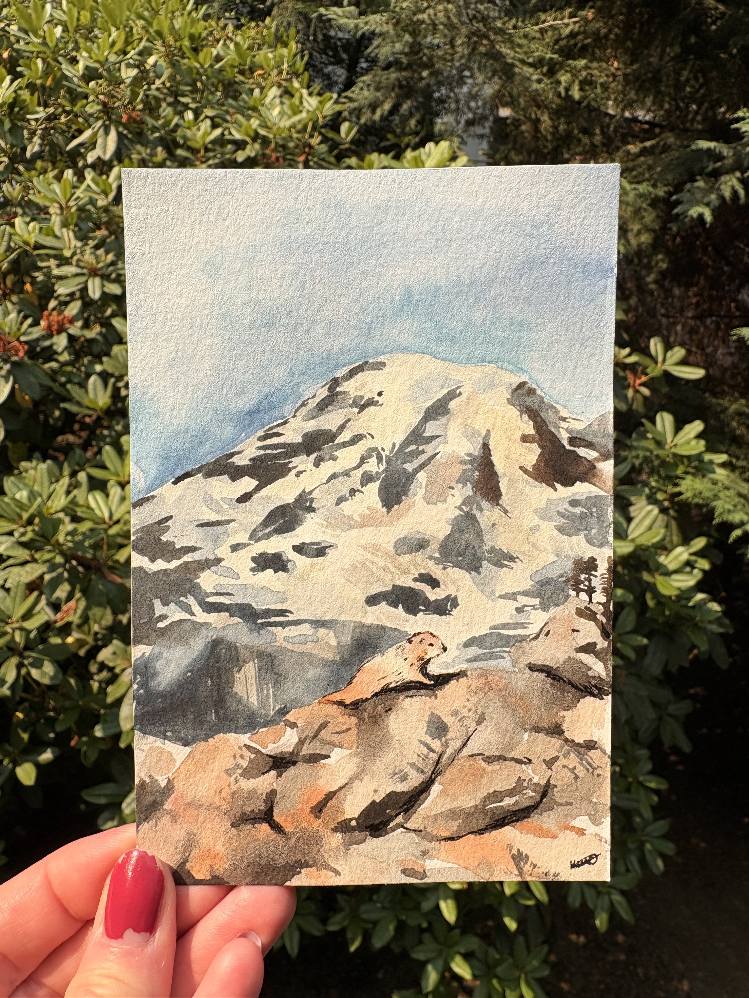 A watercolor painting of a mountain with snow-capped peaks, green and rocky slopes, and a blue sky. A small tree is visible at the bottom right corner. A person's hand with red-painted nails holds the painting in front of green shrubbery.