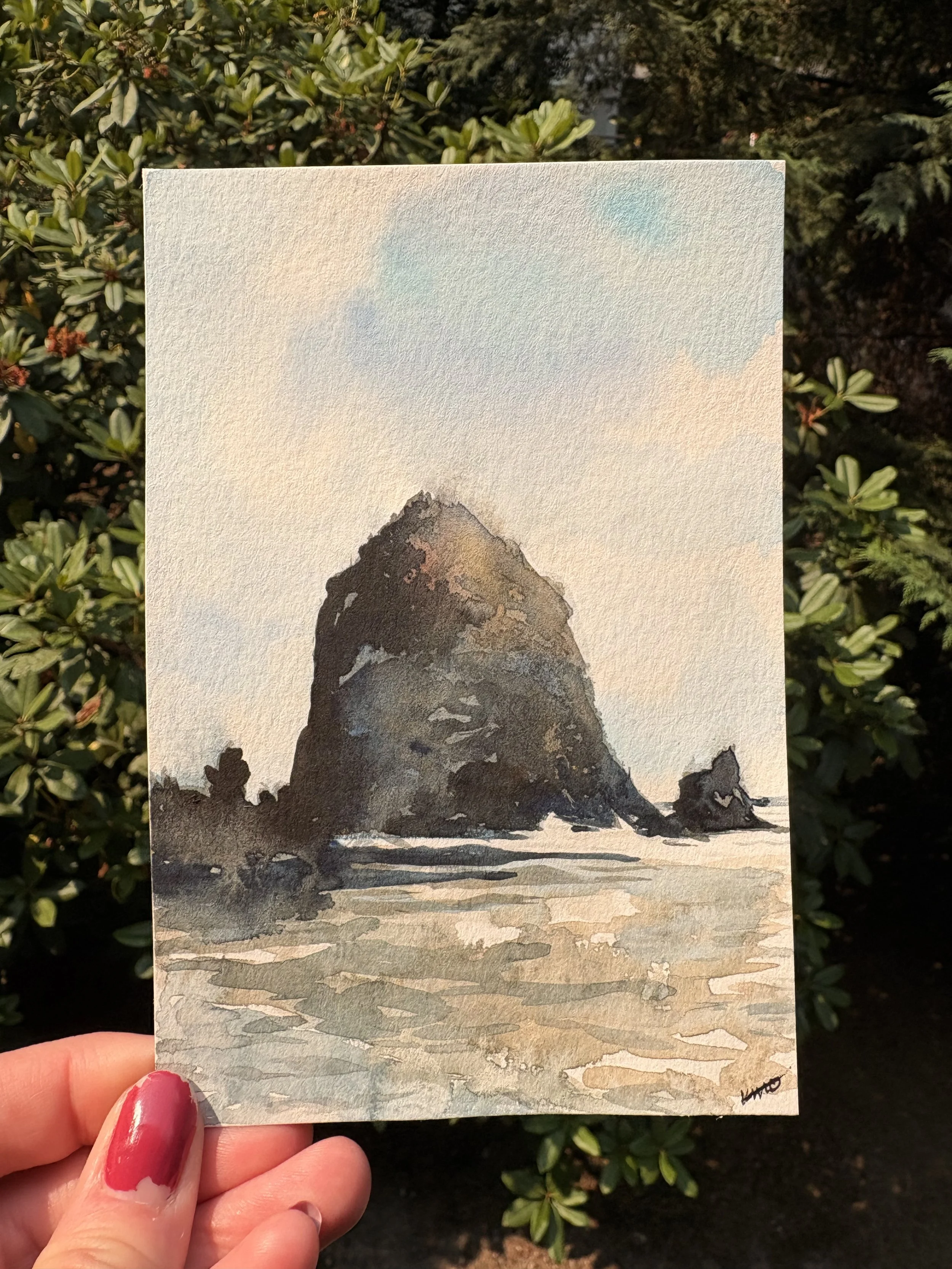 Hand holding a small watercolor painting of a mountain on textured paper, with a background of green leafy bushes.