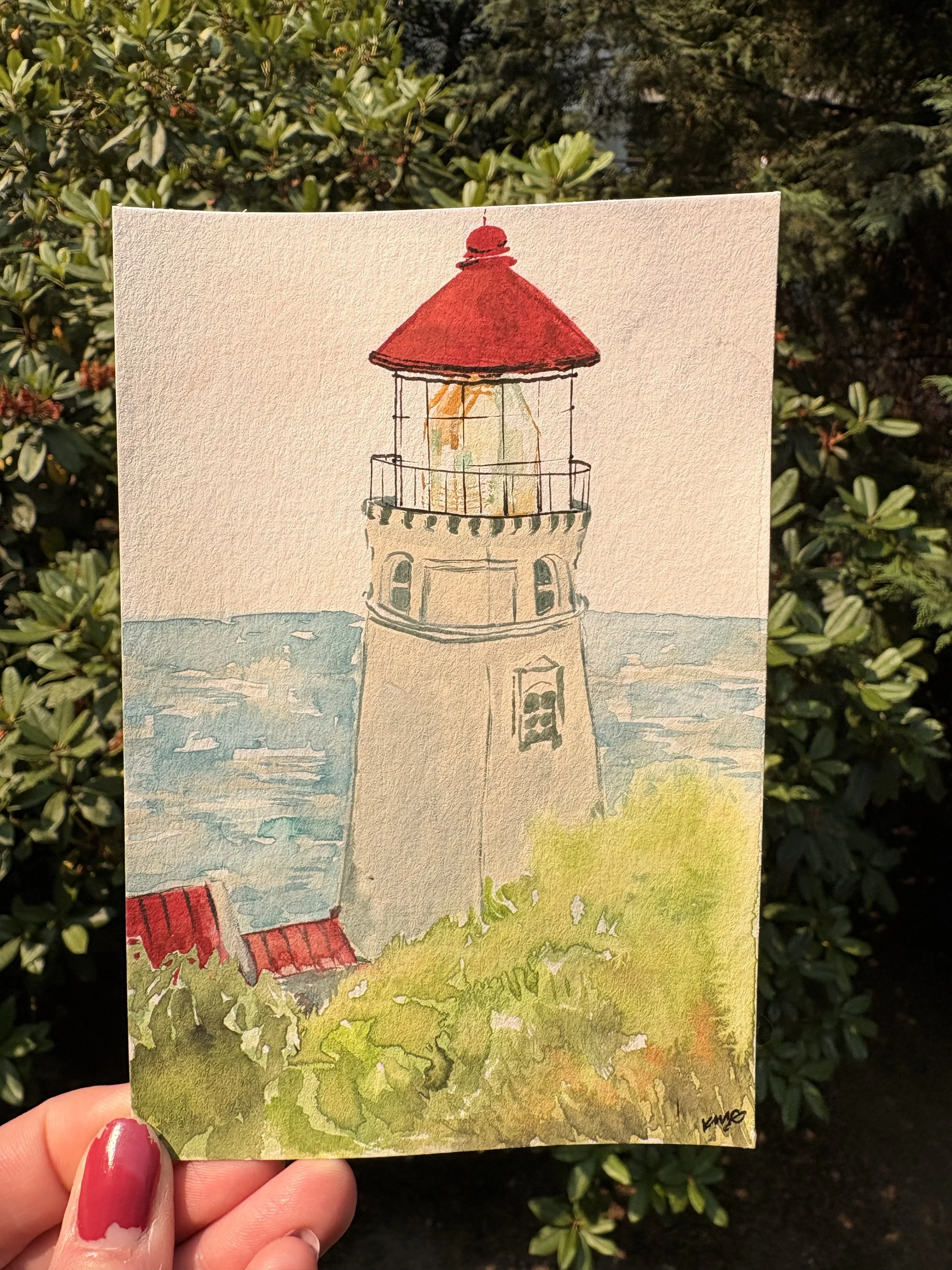 Hand holding a watercolor painting of a lighthouse with a red roof, overlooking the sea, with a grassy area at the bottom and green bushes in the background.