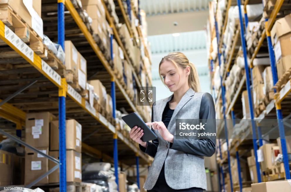 A woman in a grey blazer with black leather sleeves checks her tablet in a warehouse surrounded by tall metal shelves filled with boxes.