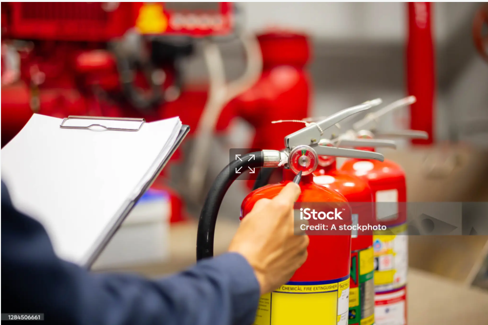 Person inspecting a fire extinguisher in a fire safety equipment room, with several red fire extinguishers in the background.
