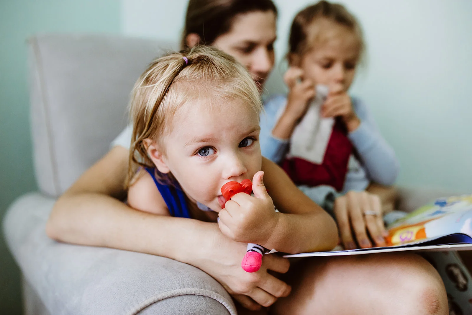 Toddler looks into camera as mom reads book to her sister