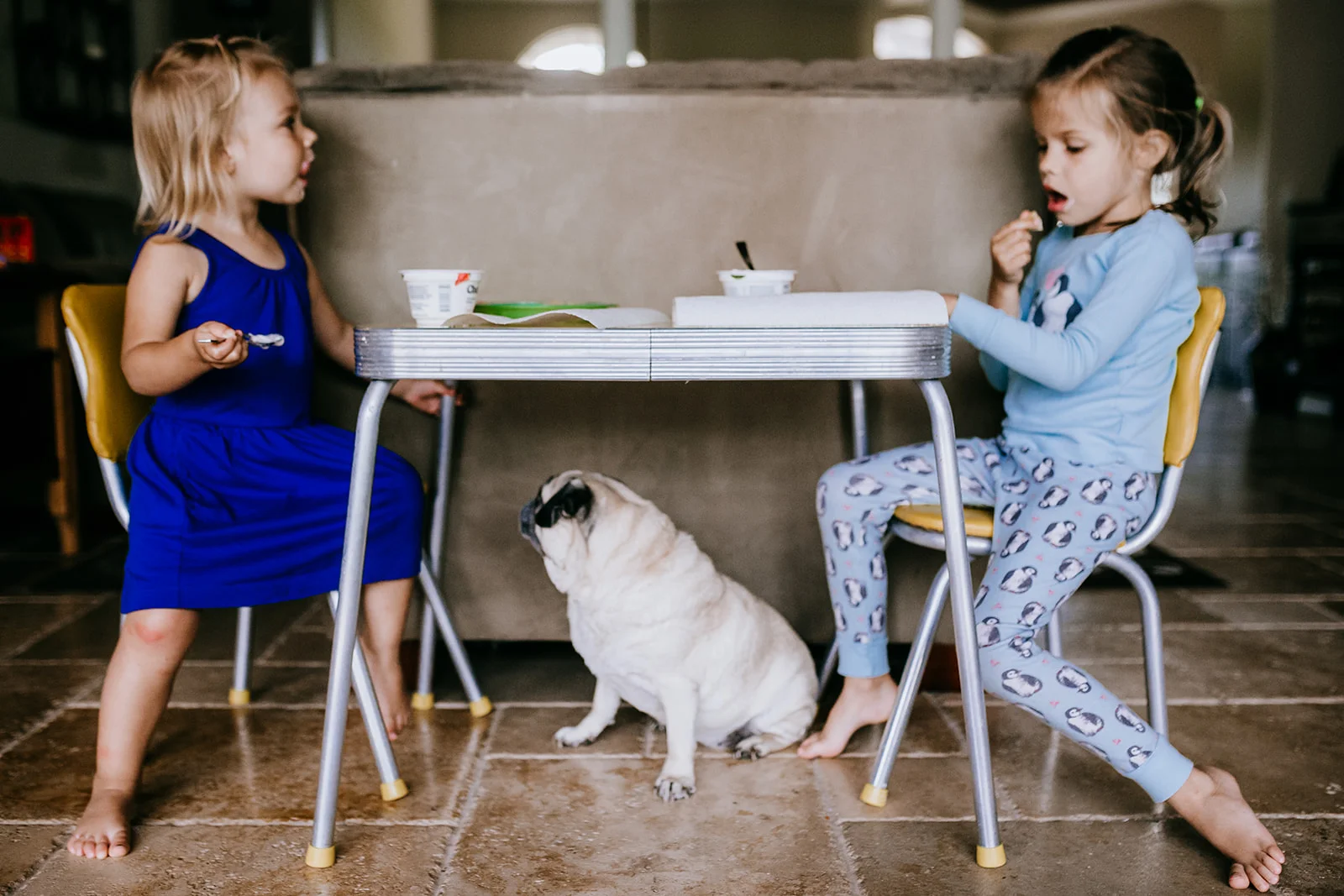 Sisters eat lunch at their kid's table while pug waits underneath for scraps