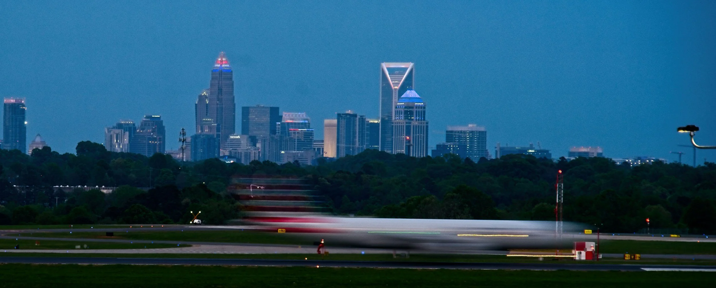 Nighttime city skyline with tall buildings illuminated, view from an airport runway with a blurred airplane taking off in the foreground.