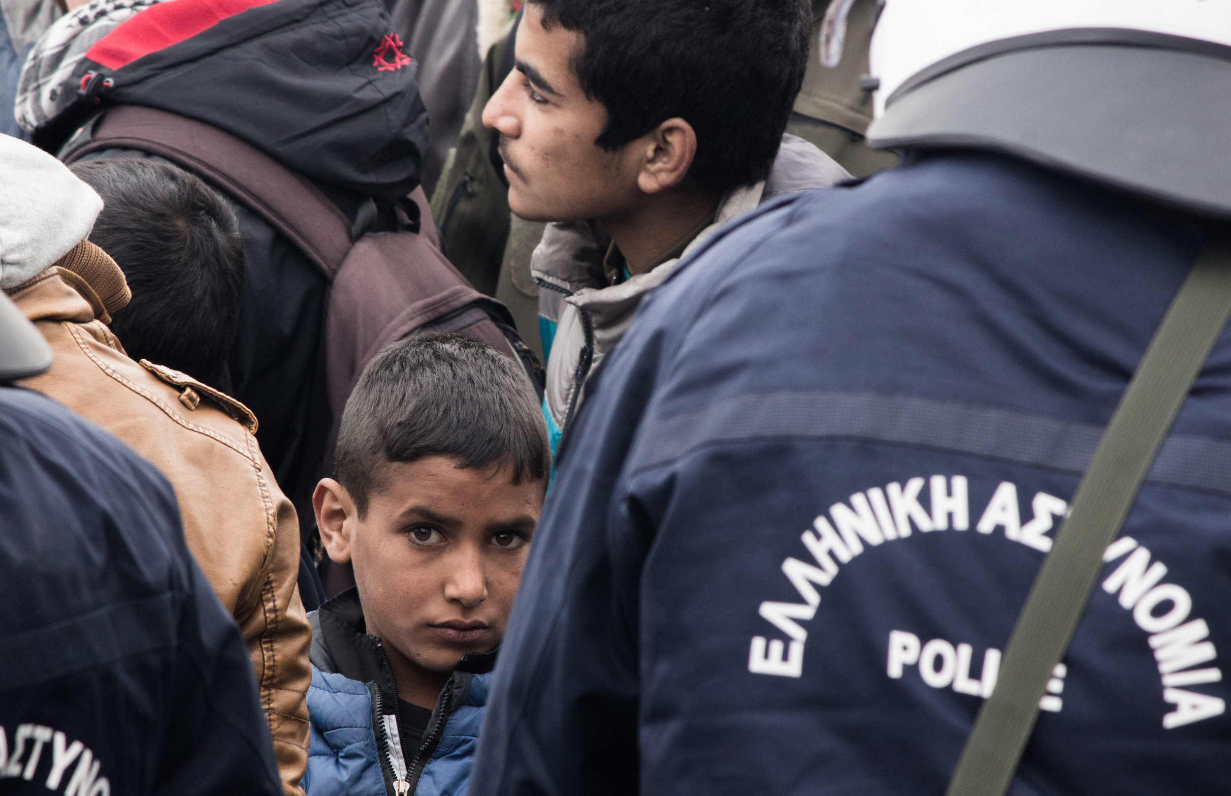 A boy looks over the shoulder of Greek riot police during a large protest in the border camp of Idomeni, March 2016. Tensions had been mounting for several days and ultimately a large, but peaceful, protest was held with several hundred people in at