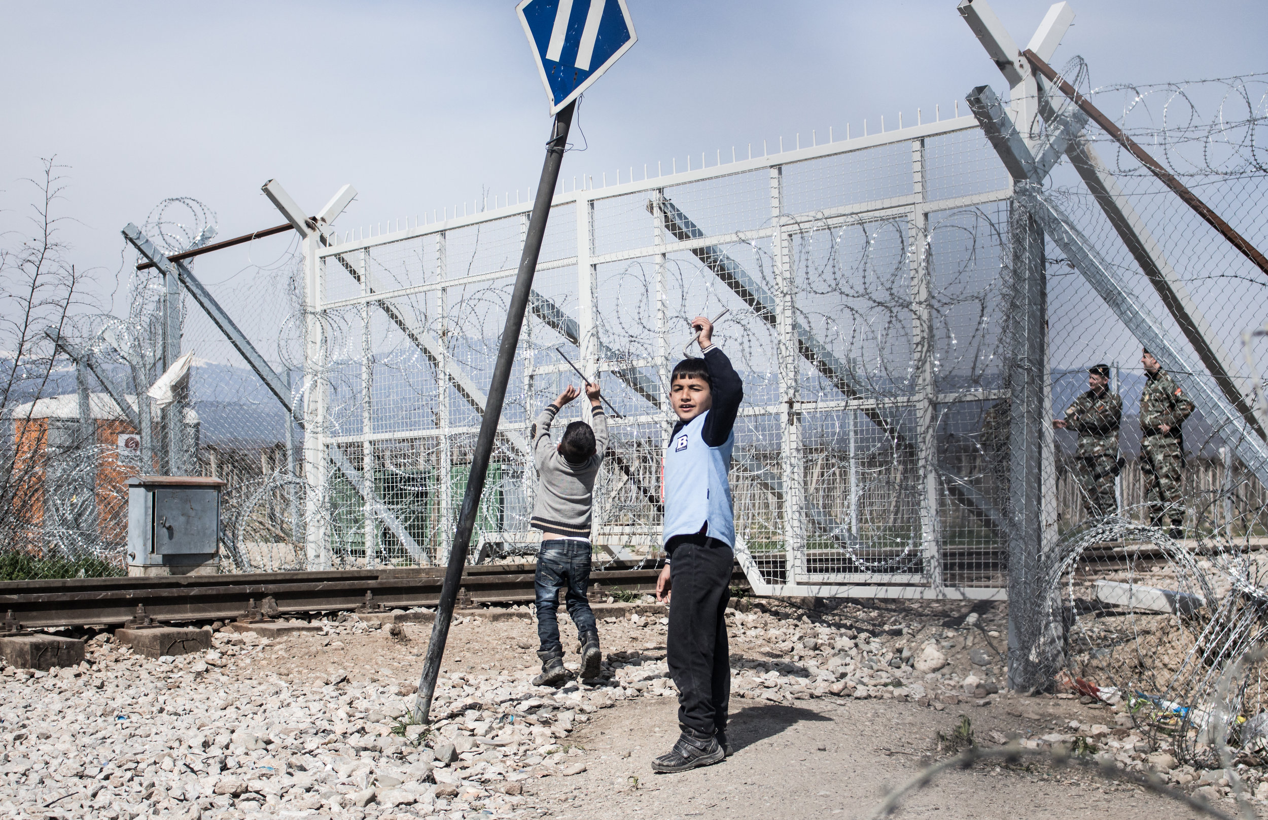 Two boys playing/ protesting by the railway crossing point of the Greek-FYROM border, while Macedonian guards look on across the fence, March 2016. 