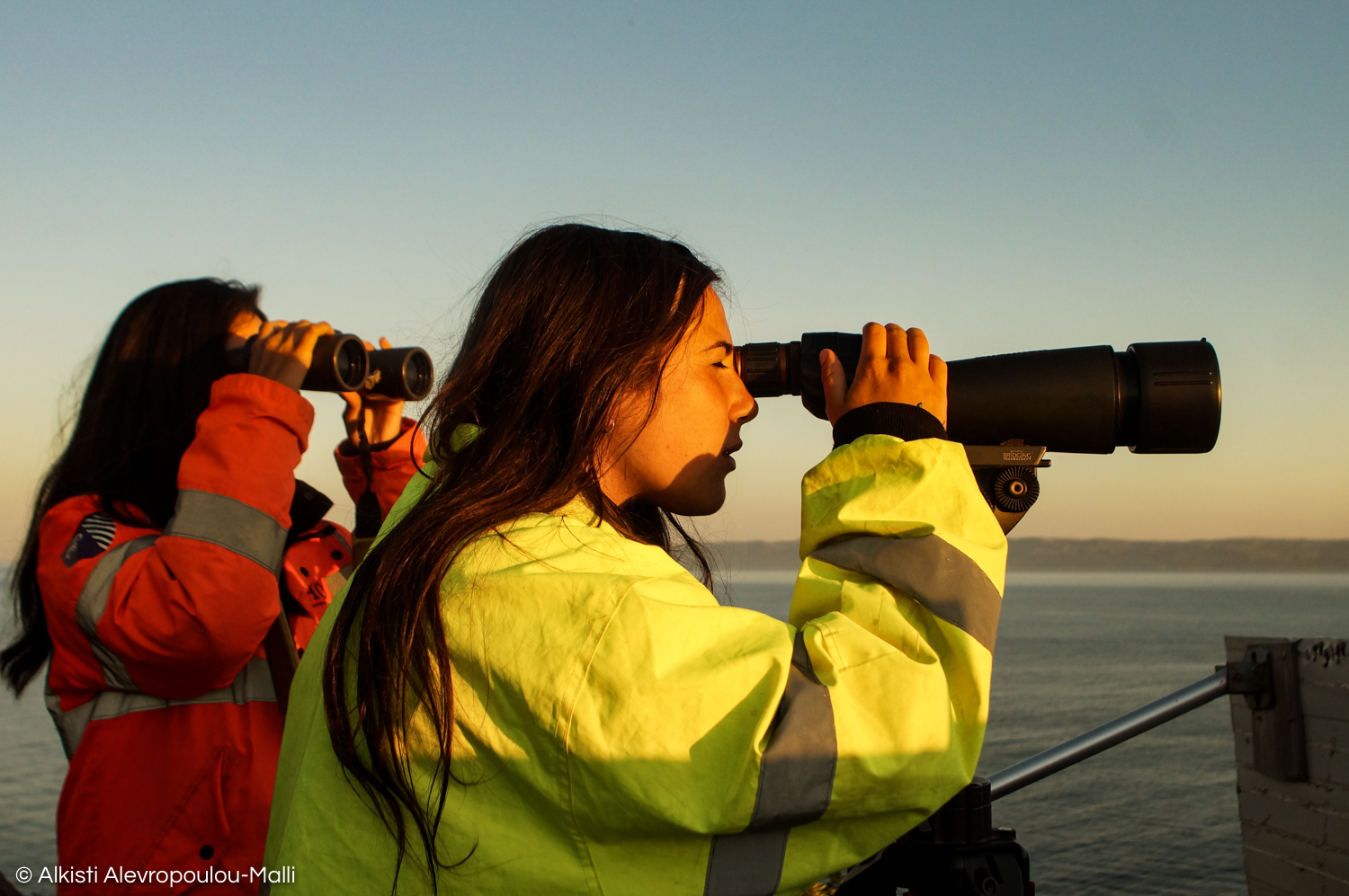 Sunrise in Lesvos, and two volunteers scan the sea with binoculars and a telescope for potential boats crossing over from Turkey.