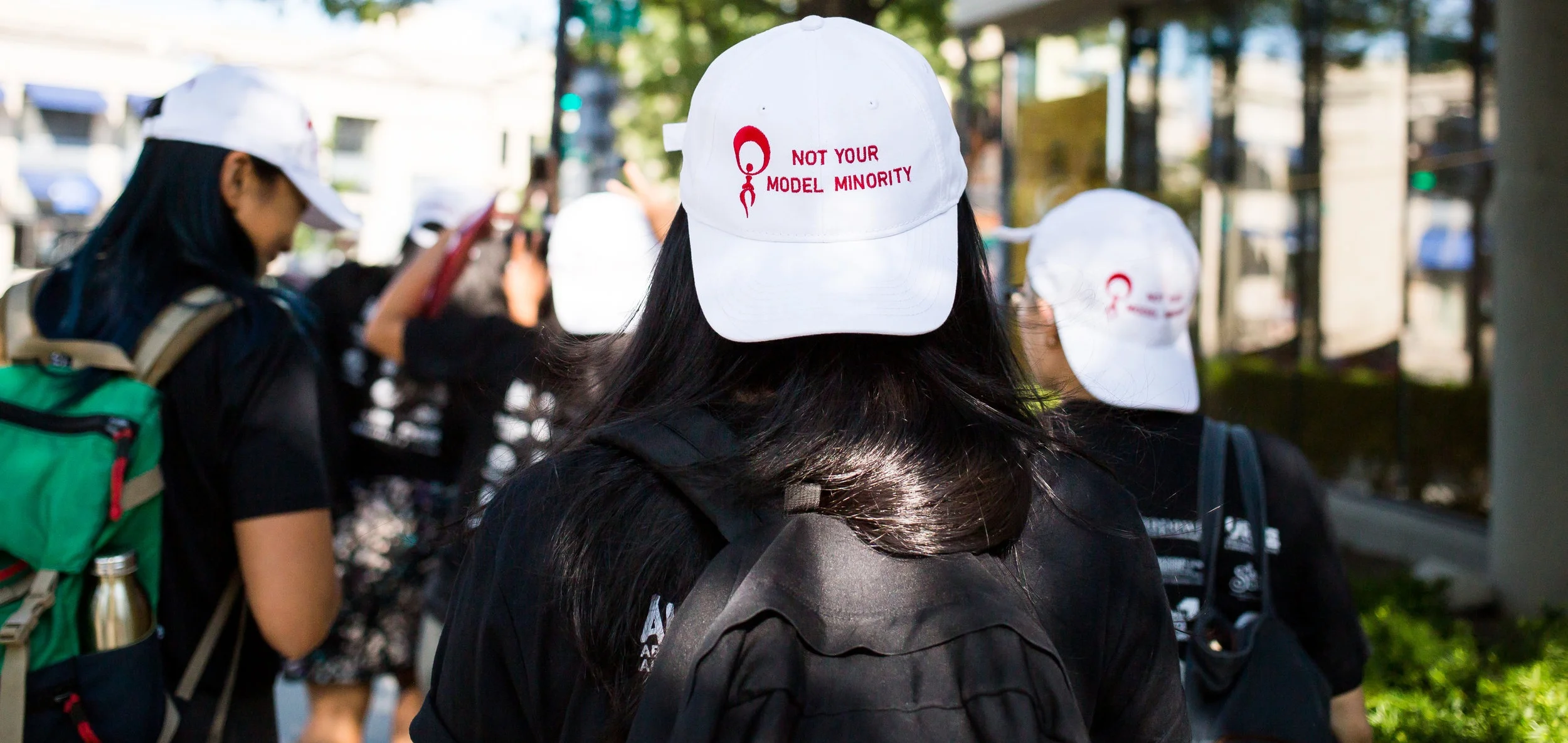 A person with flowing black hair is wearing a cap that reads “NOT YOUR MODEL MINORITY.” The cap is worn backwards, and several other people are wearing the same hat in the background.