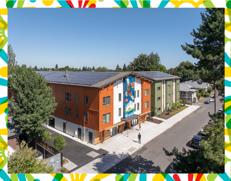 Photograph of a three-story apartment building, painted white, rust-orange, and sage-green, with a colorful mural, seen from a distance and above.