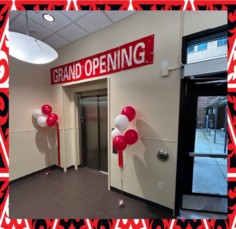 Photograph of an elevator entrance with a red and white "Grand Opening" banner above the doors and clusters of red and white balloons on either side.
