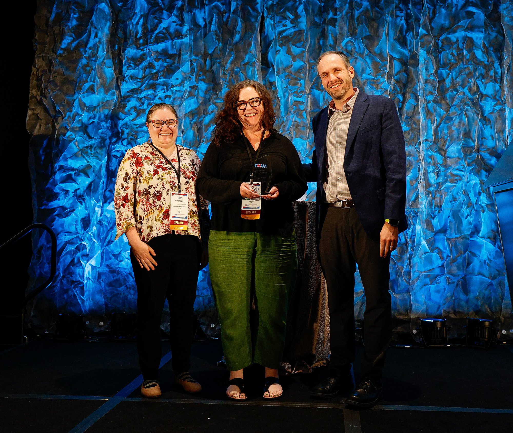 Photograph of three people on stage with blue backdrop, after award acceptance