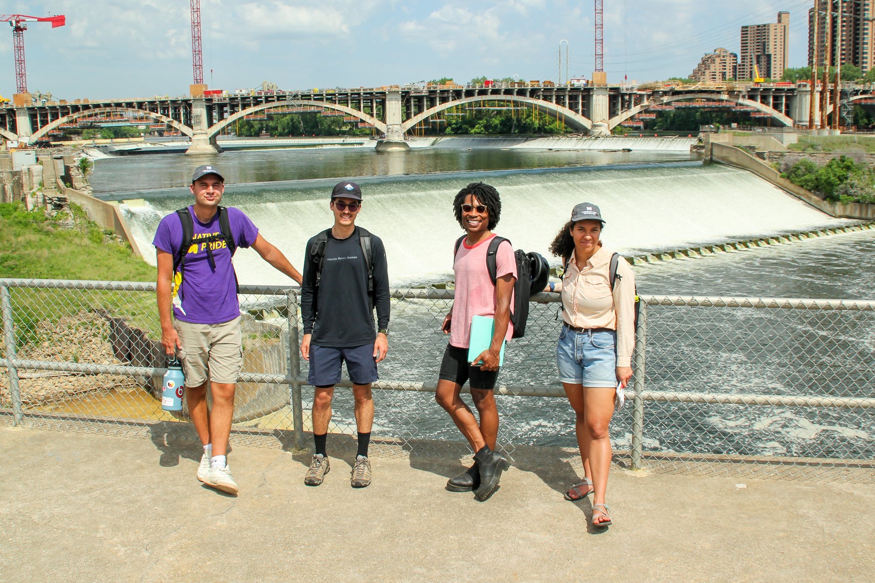  Four people stand against the railing at the Upper St. Anthony Falls Lock and Dam with the falls and a large bridge behind them in the background. 