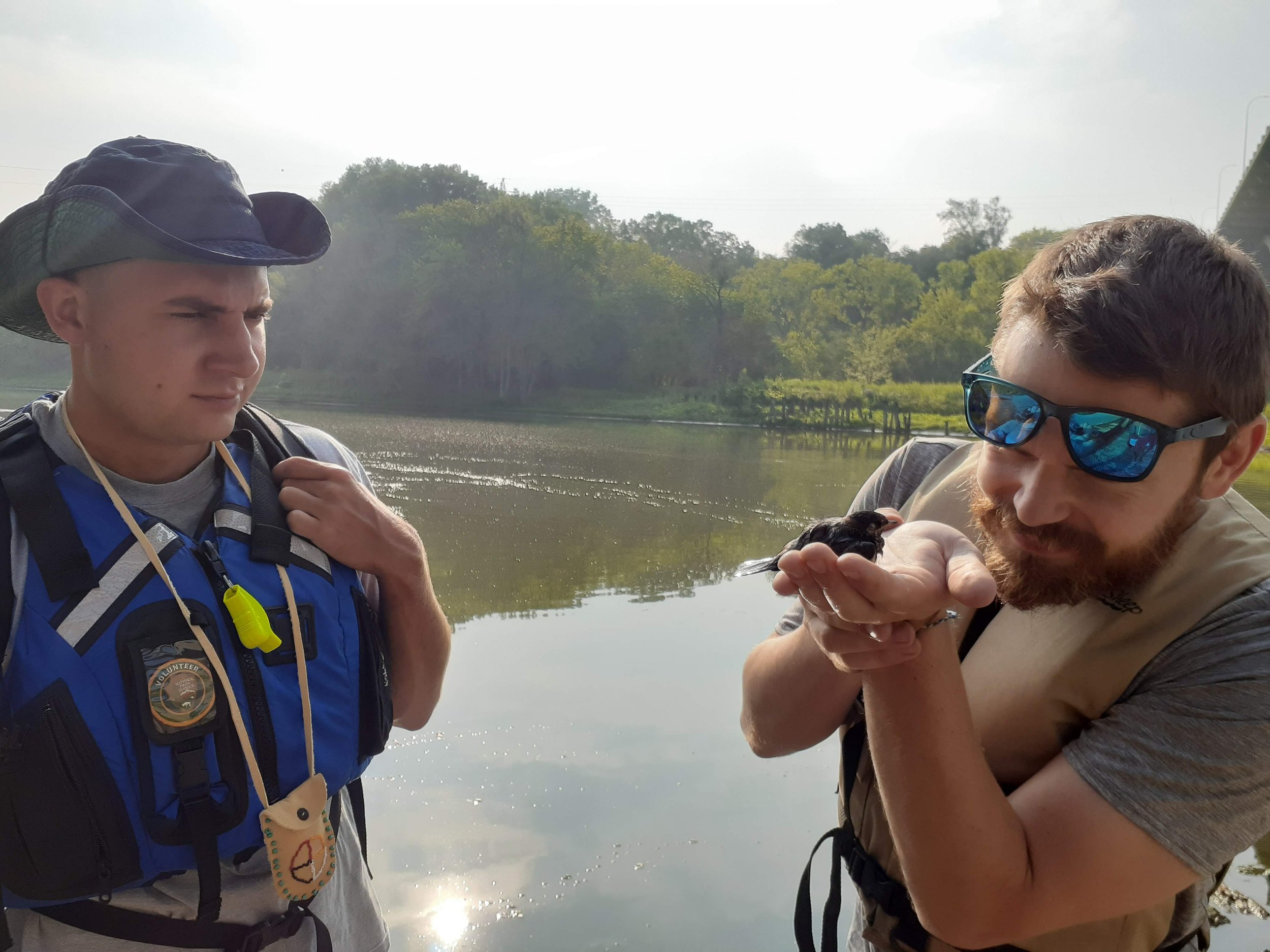  A person holds a bird in his hand while another person watches. They are both wearing life jackets and standing near water. 