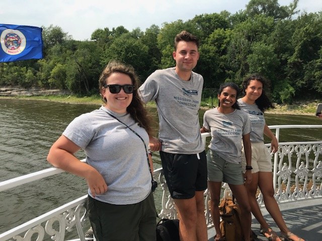  Four people stand against the railing on the deck of a boat on the Mississippi River. They are smiling and looking at the camera. 