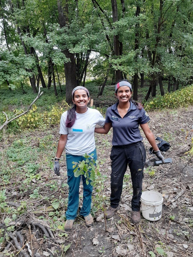  Tracy Few and Tamara Few  Two people stand in the woods. They have just planted some trees and are standing next to a large white water bucket.  