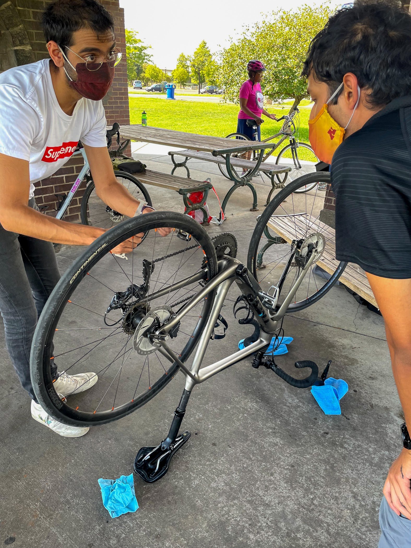  Pallav Kumar (left)  A person demonstrates how to fix a bike part outside under an outdoor pavilion. The bike is flipped upside down and another person watches. 