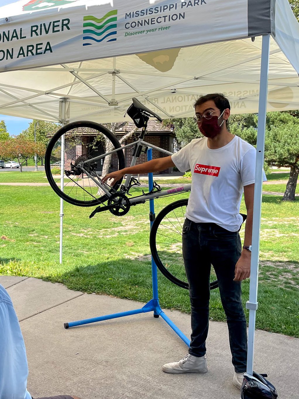  Pallav Kumar  A person demonstrates in front of a bike propped up in a bike stand. They are standing under a white canopy tent outdoors. 